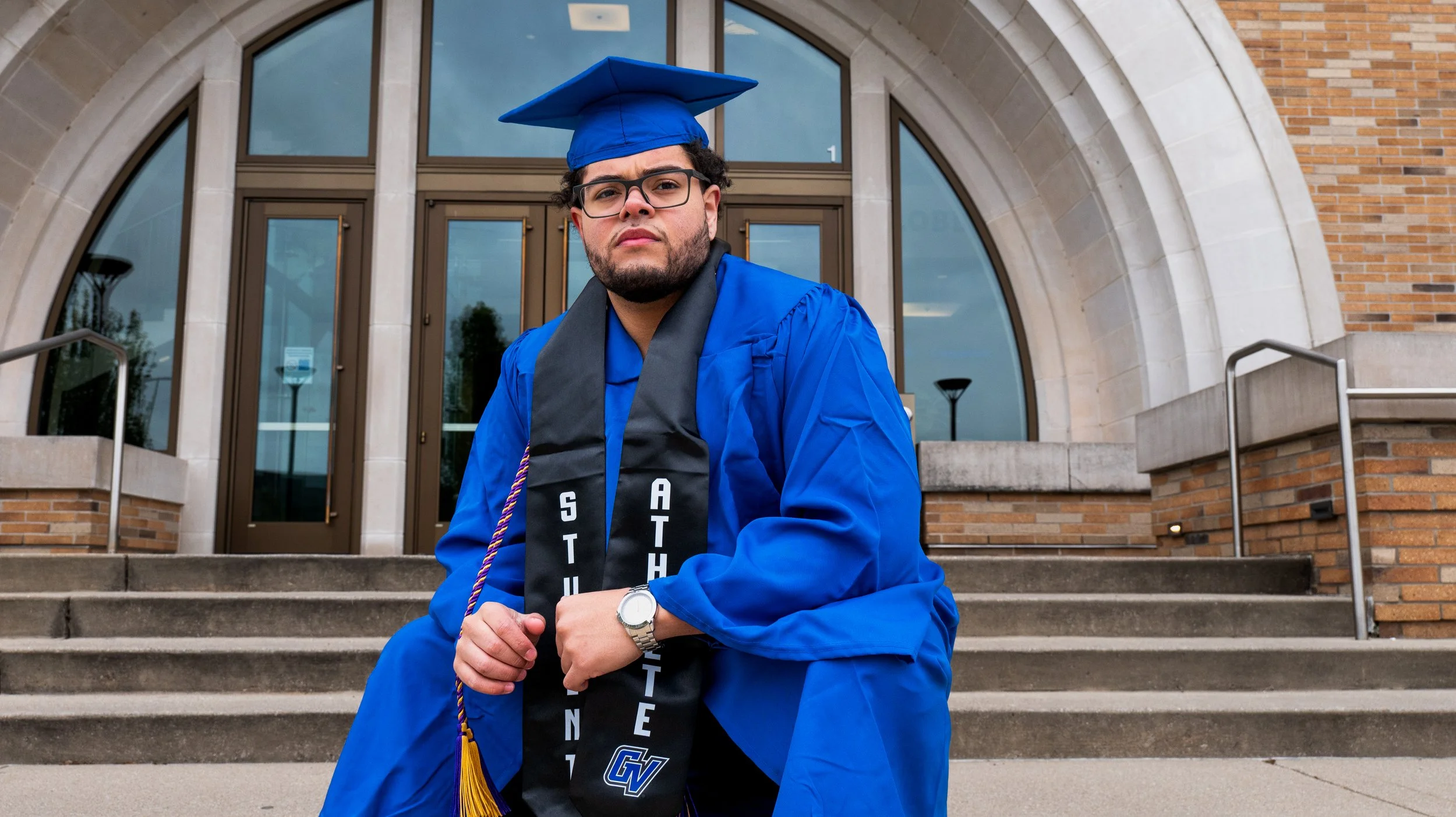 A young man wearing glasses and a blue graduation gown and cap sitting on steps in front of a building with large windows and brick walls.