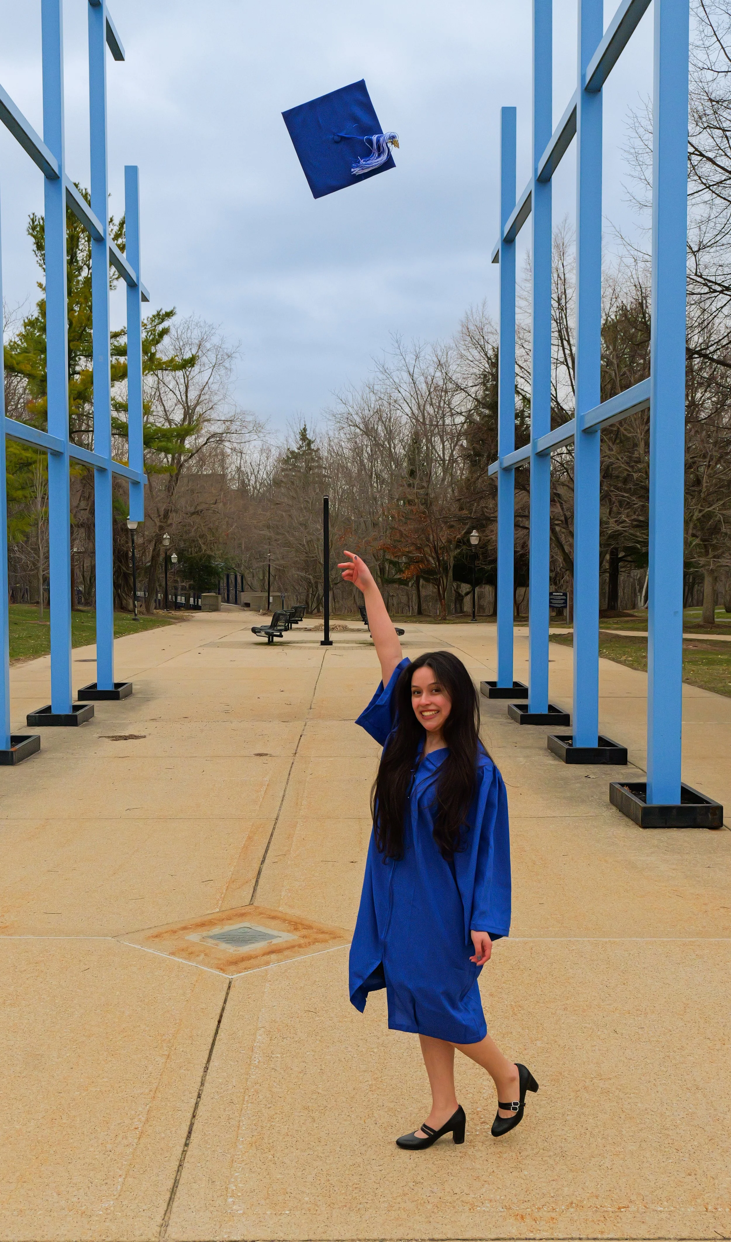 A young woman in a blue graduation gown and heels stands outdoors on a paved walkway, smiling and pointing at her floating graduation cap. The cap is mid-air above her head against a cloudy sky, with trees and empty benches in the background.