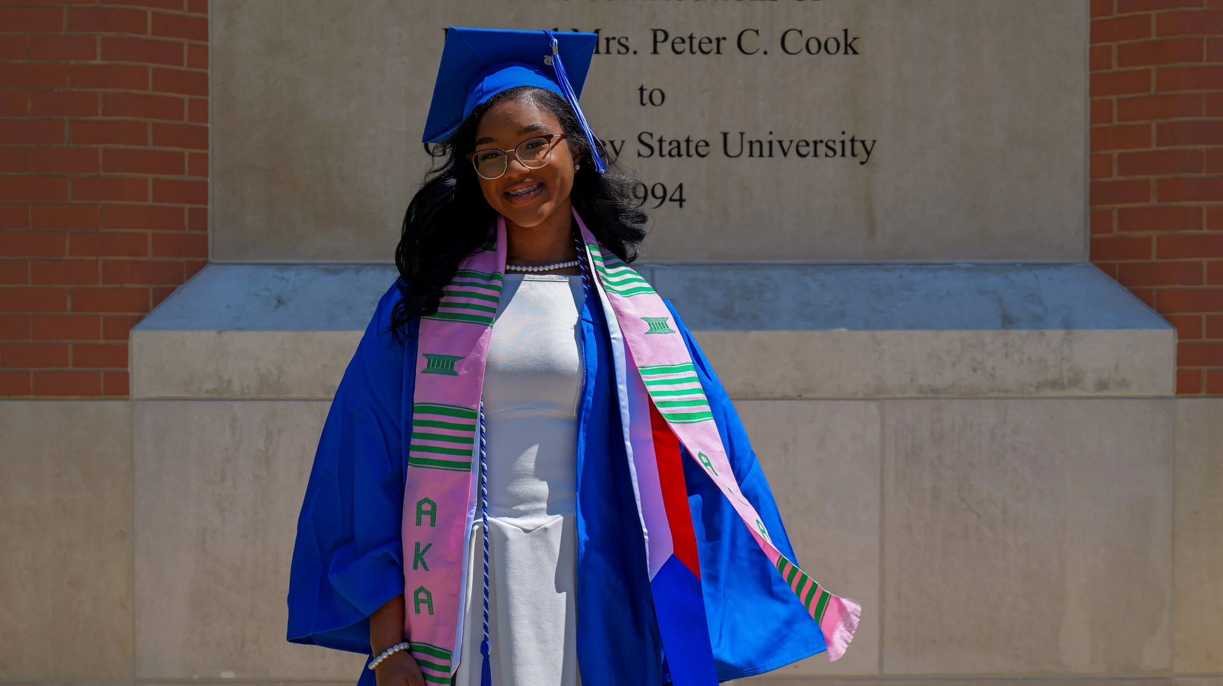 A young woman in a blue graduation cap and gown standing in front of a university sign. She is smiling and wearing glasses and a colorful sash with green and purple patterns.