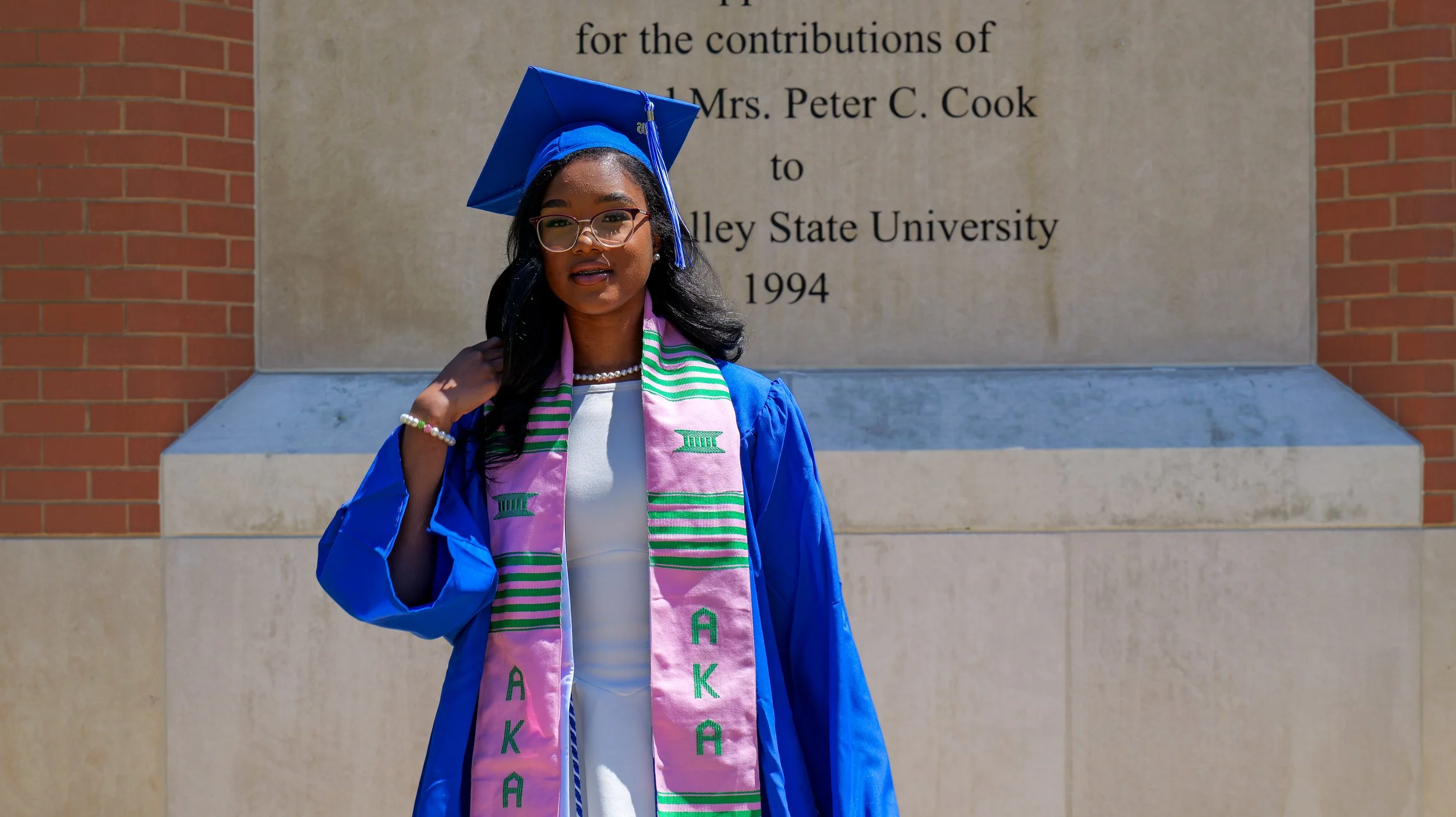 A young woman in a blue graduation gown and cap standing in front of a stone wall with an inscribed plaque. She is wearing glasses, a pearl necklace, and a pink stole with green lettering.