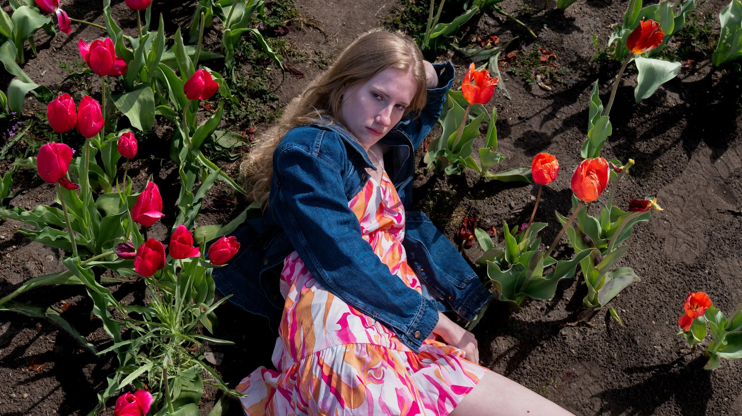 A young woman with long, wavy blonde hair sits on the ground in a flower bed with pink and orange tulips, wearing a colorful dress and a denim jacket.