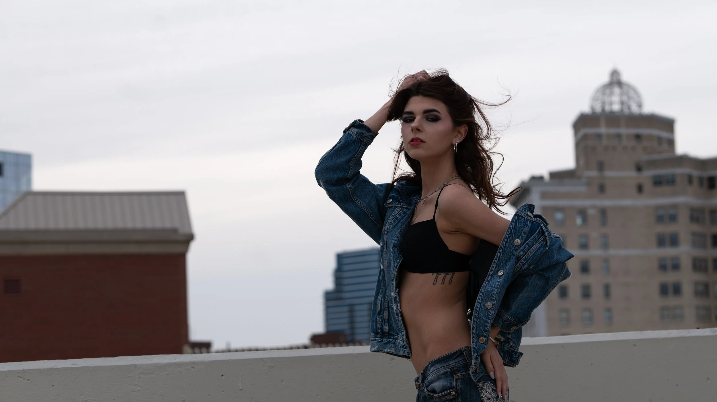 A woman with dark hair and makeup poses on a rooftop with city buildings in the background. She is wearing a black top, a denim jacket, and jeans, and has tattoos visible on her side and arm.