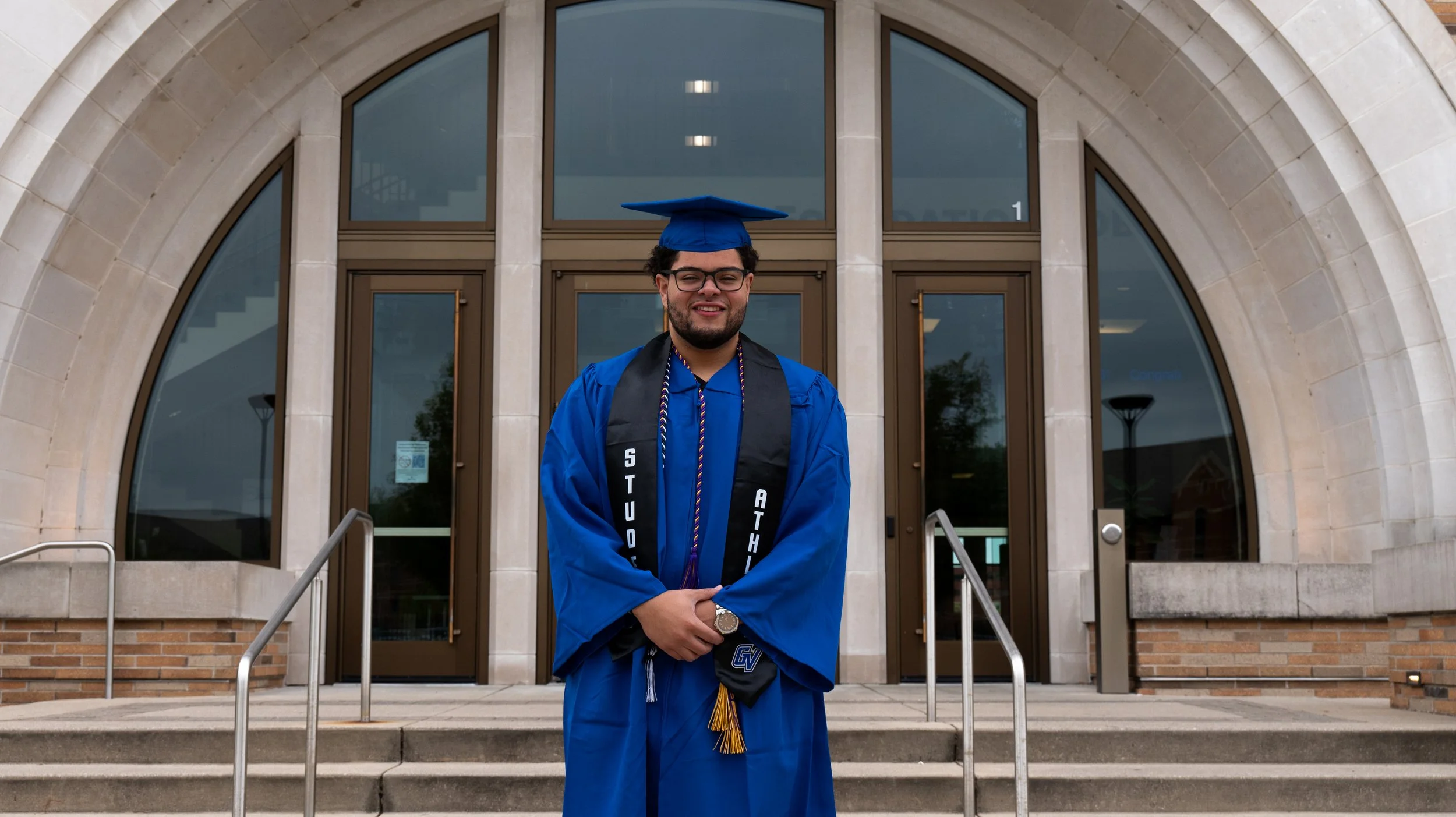 A young man in a blue graduation gown and cap standing in front of a building with arched windows, smiling with his arms crossed.
