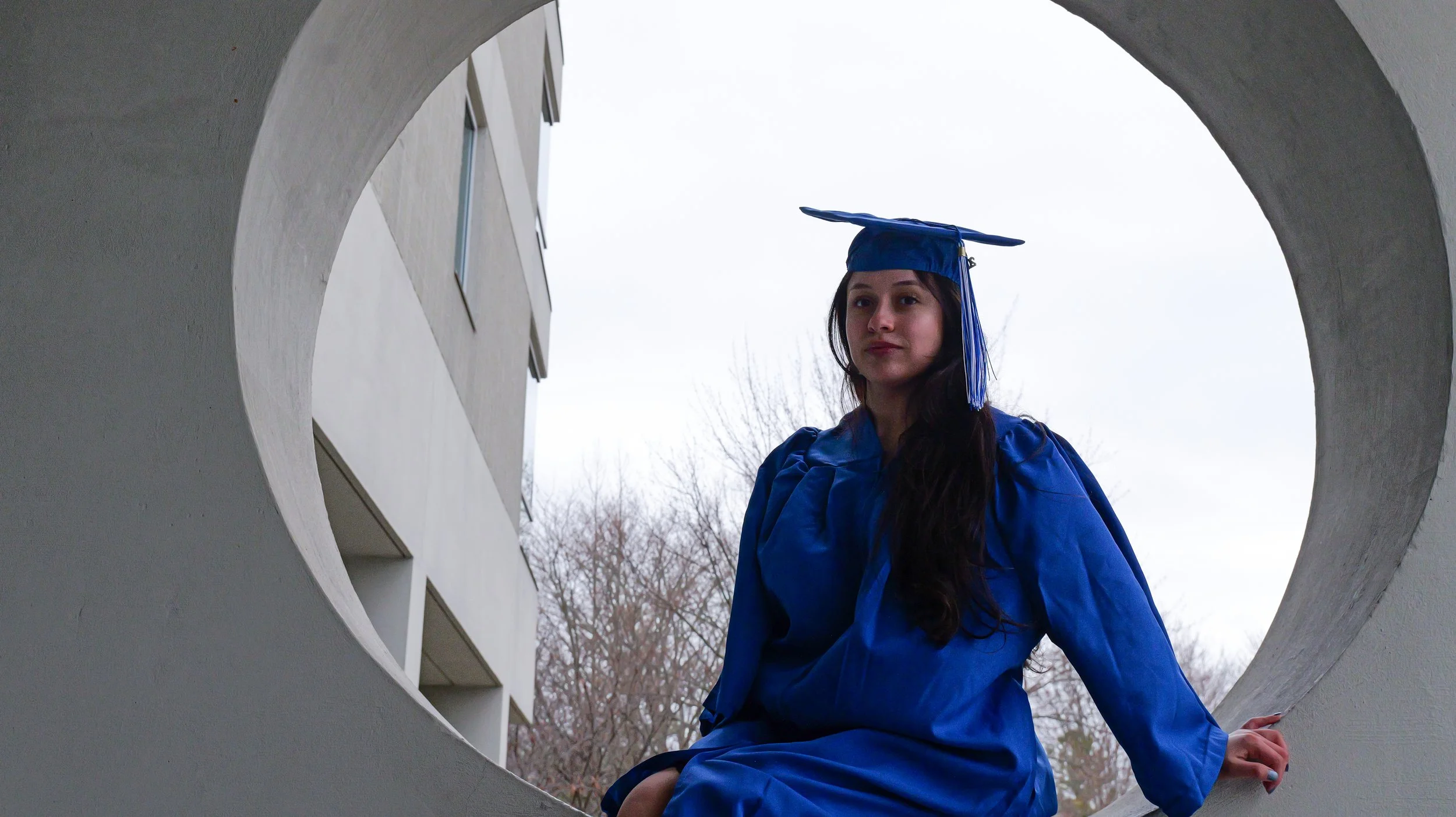 A woman dressed in a blue graduation gown and cap sitting on a concrete structure, framed by a circular opening, with a building and leafless trees in the background, overcast sky.
