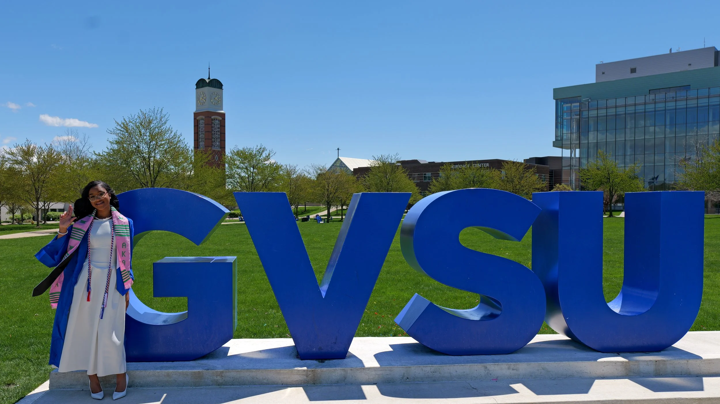 A woman in a white dress and graduation gown standing in front of large blue letters spelling 'CVSU' in an outdoor setting with green grass, trees, a clock tower, and modern buildings in the background on a sunny day.