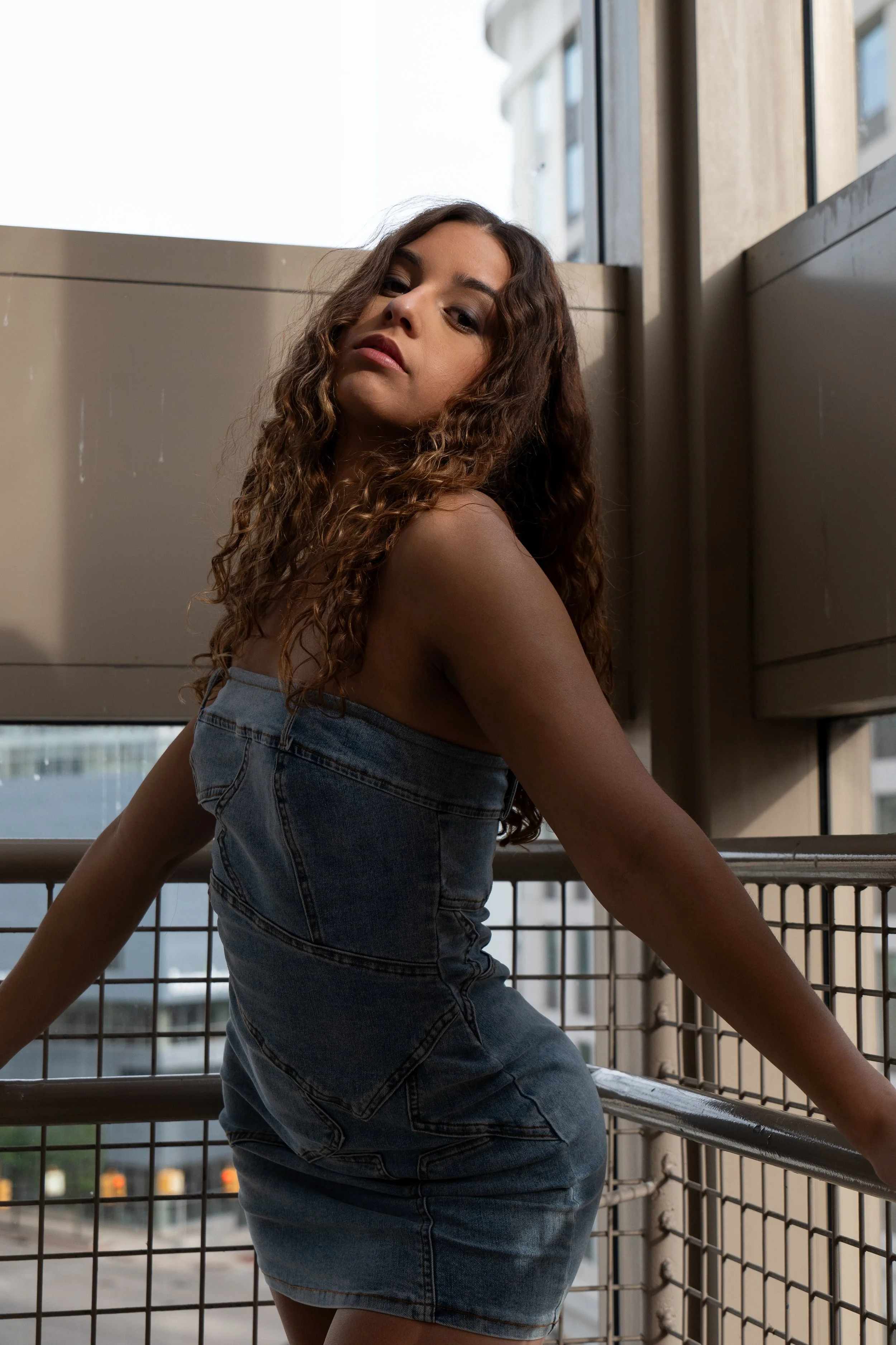 Young woman with long curly hair wearing a denim dress, leaning against a metal railing in an urban outdoor setting, with buildings and a street in the background.