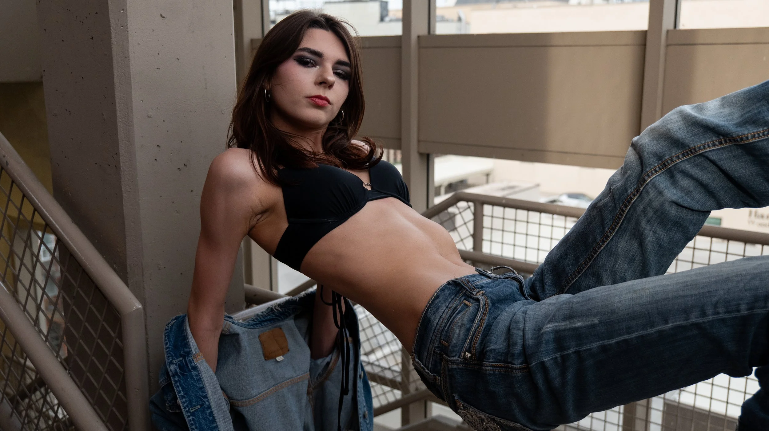 A woman with dark hair and makeup lying on a balcony railing, wearing a black top and jeans, with an urban cityscape in the background.