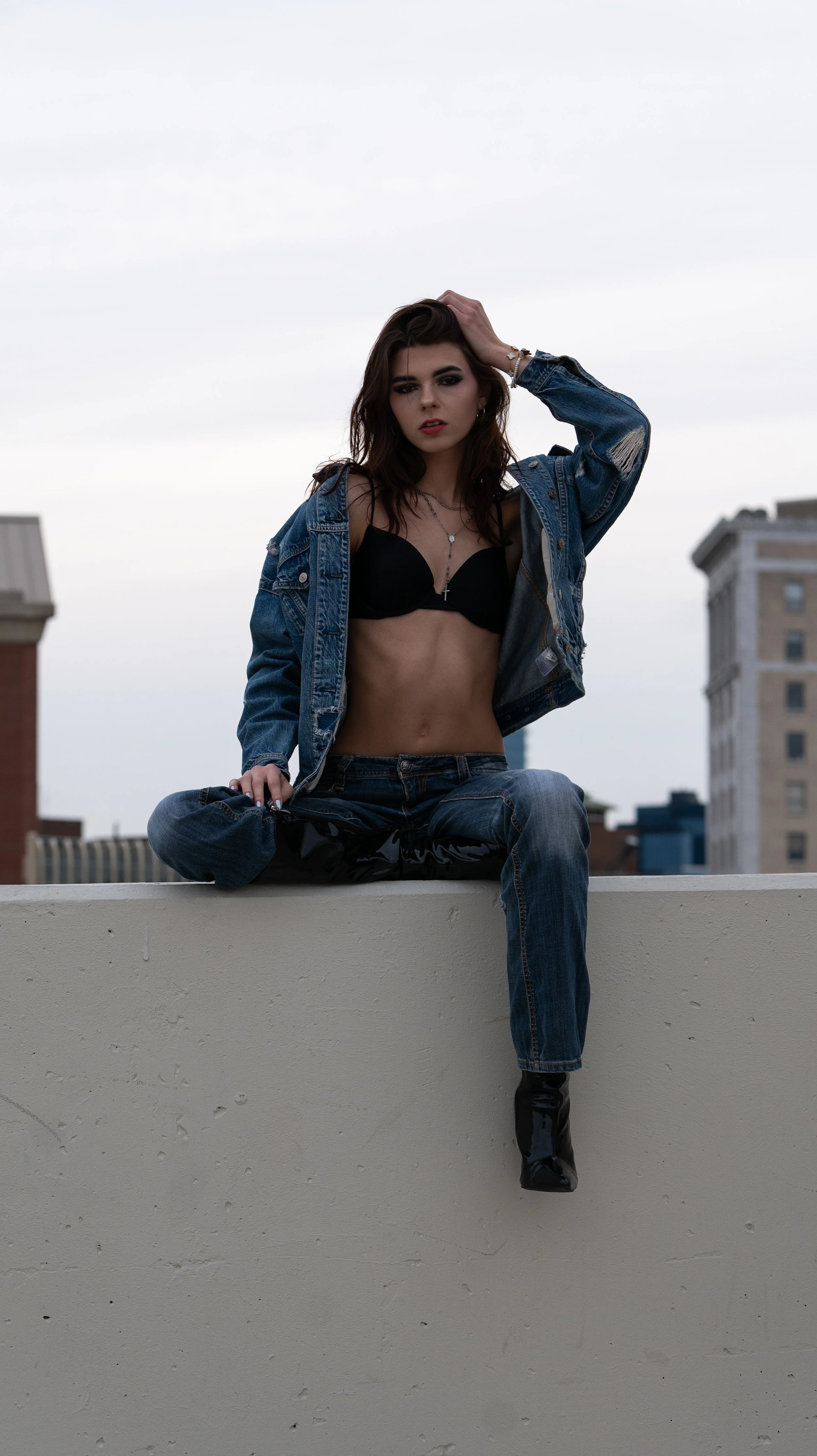 A young woman with dark hair, black makeup, and a confident expression is sitting on a rooftop ledge with her legs crossed. She is wearing a black bra, a blue denim jacket, ripped jeans, and shiny black boots. The city skyline is visible in the backg