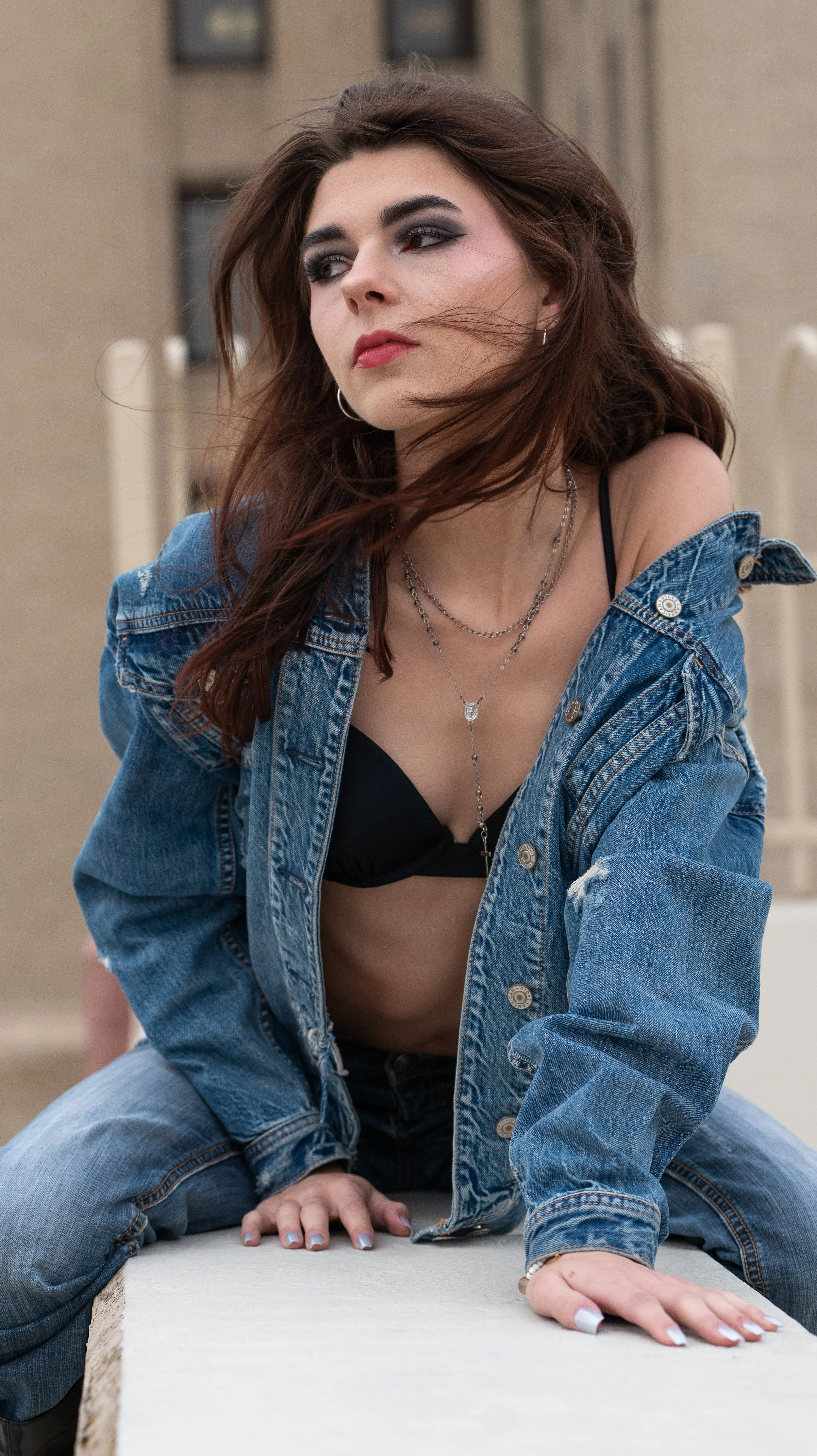 A woman with long brown hair, wearing a denim jacket slightly off her shoulders over a black bra, layered necklaces, and hoop earrings, is sitting on a beige surface with her hands on it, outdoors with blurred city buildings in the background.