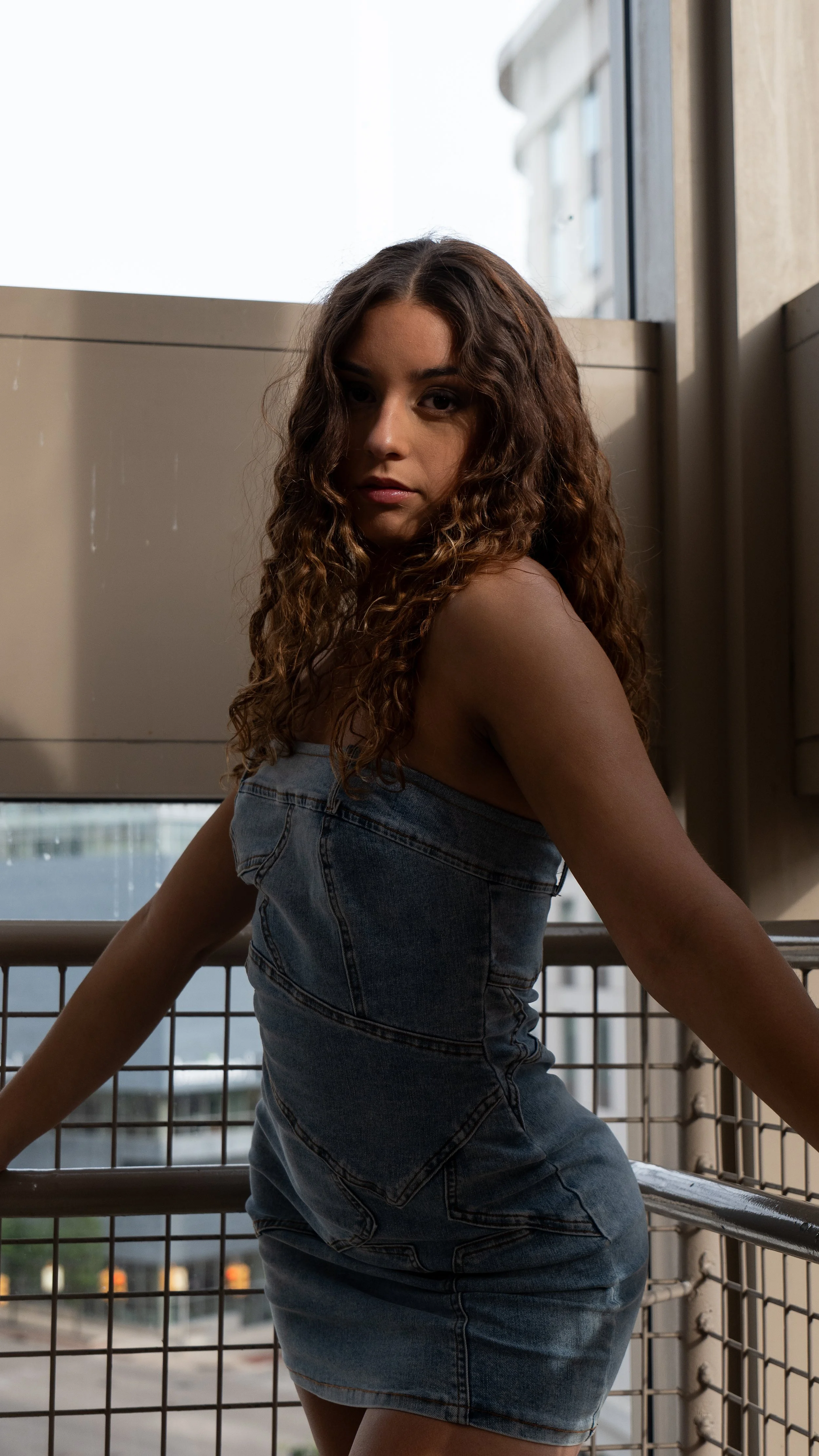A woman with curly brown hair wearing a denim dress on an outdoor balcony