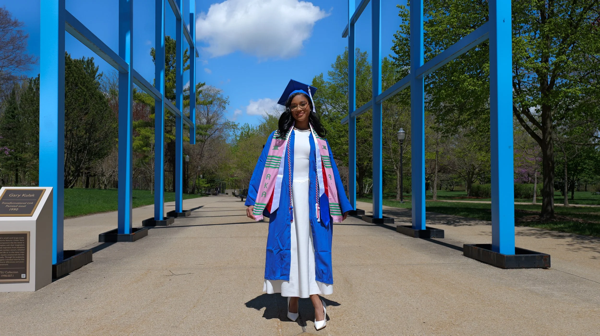 Woman in white dress and blue graduation gown standing under a modern blue steel installation in a park with trees and blue sky.