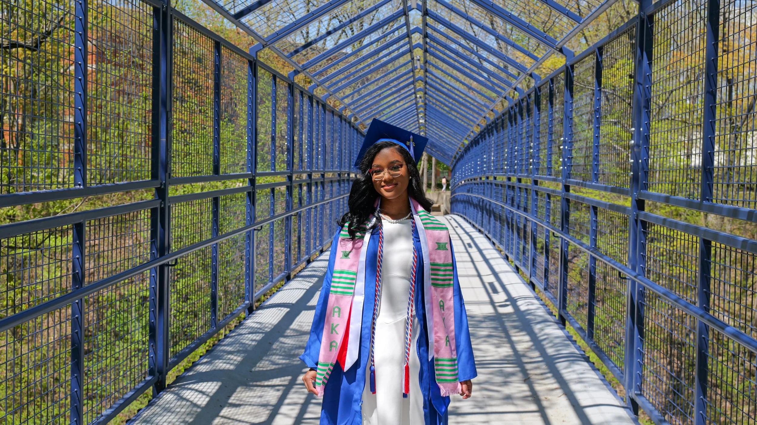 A young woman in a graduation cap and gown standing on a blue bridge, with a colorful scarf draped around her neck, outdoors with trees in the background.