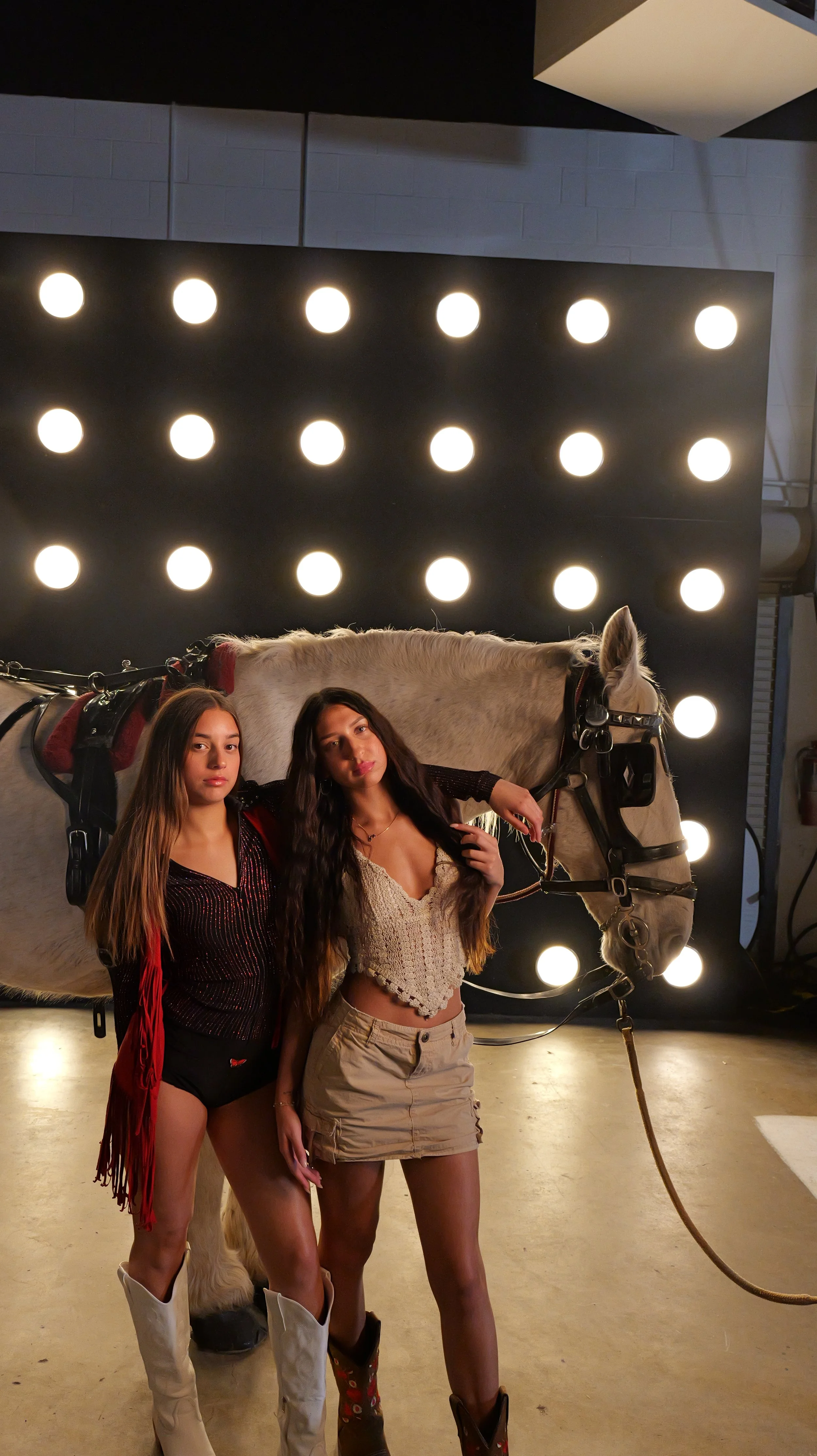 Two young women posing with a white horse in an indoor space with a black wall and bright circular lights overhead. One woman has long brunette hair and is wearing a black and red outfit, while the other has long brown hair and is wearing a beige cro