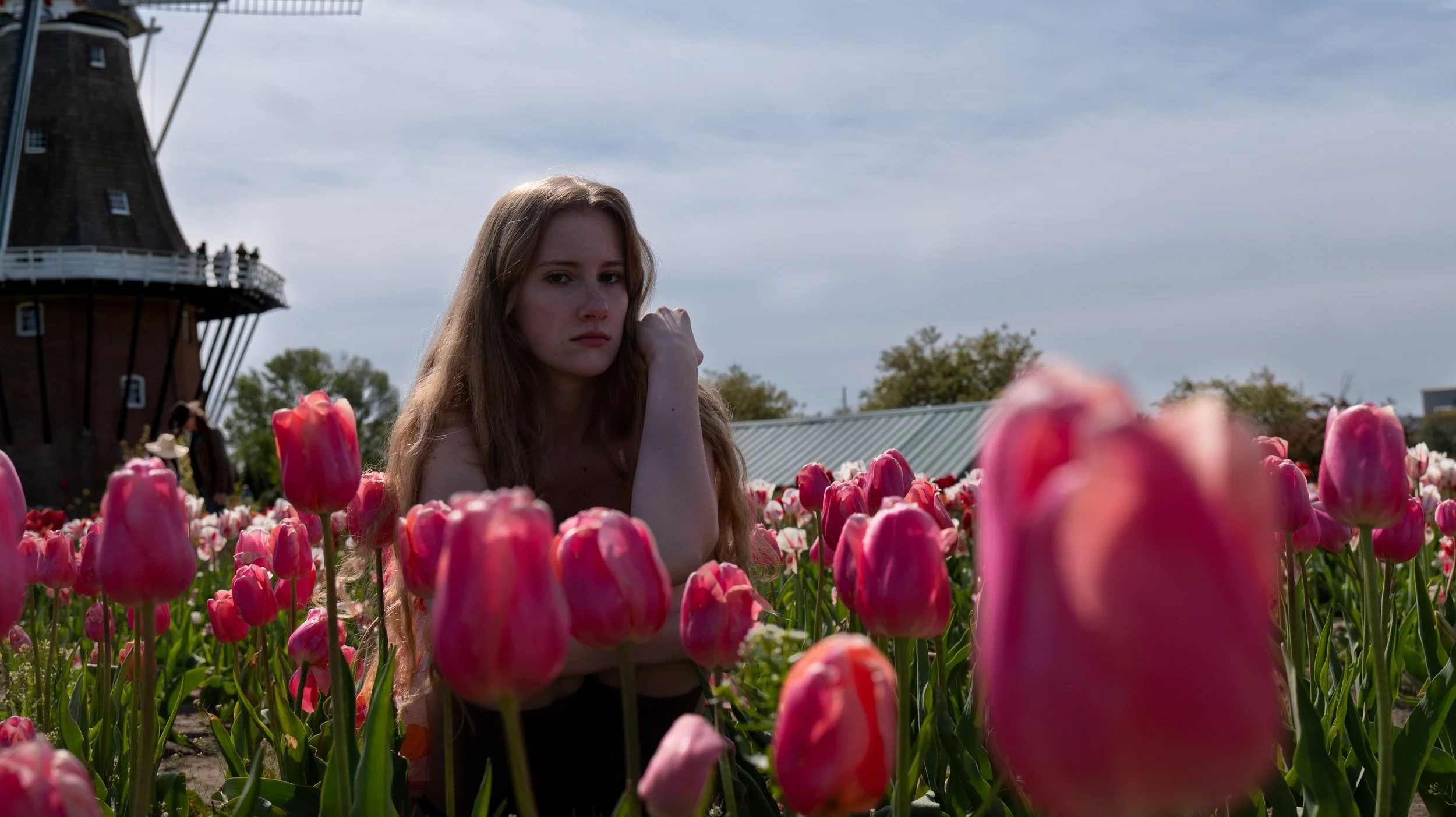A young woman with long, wavy red hair is surrounded by pink tulips in a field, with a windmill and partly cloudy sky in the background.