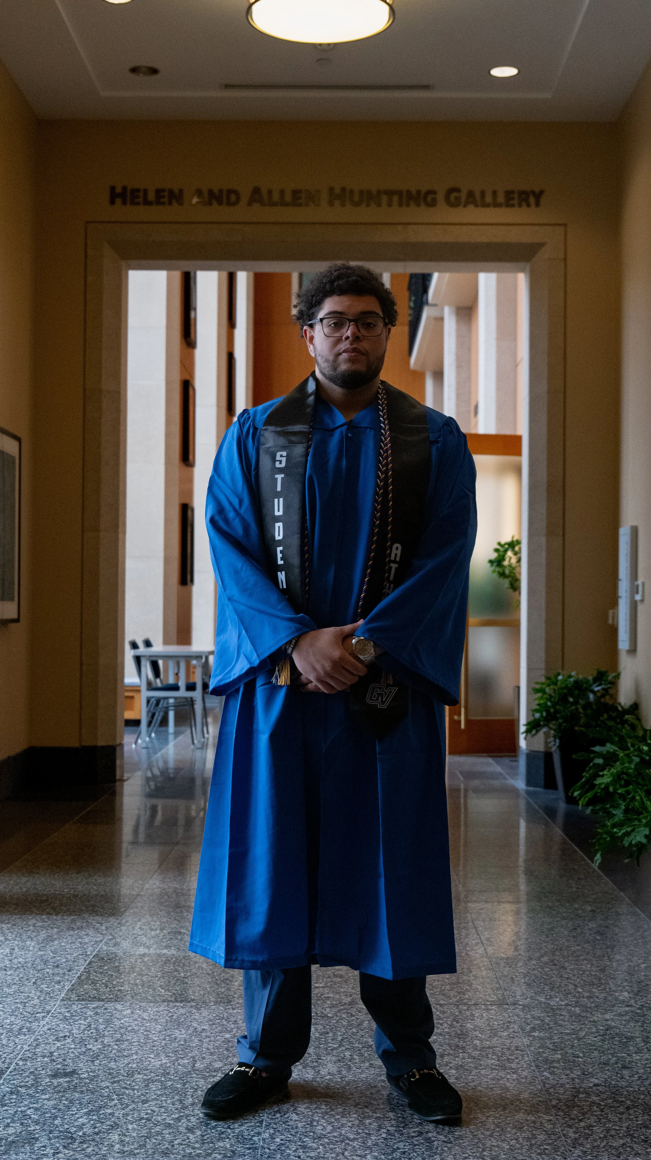 A man in a blue graduation gown and black tassel stands with arms crossed in front of a sign that reads 'Helen and Allen Hunting Gallery' in a building's interior.
