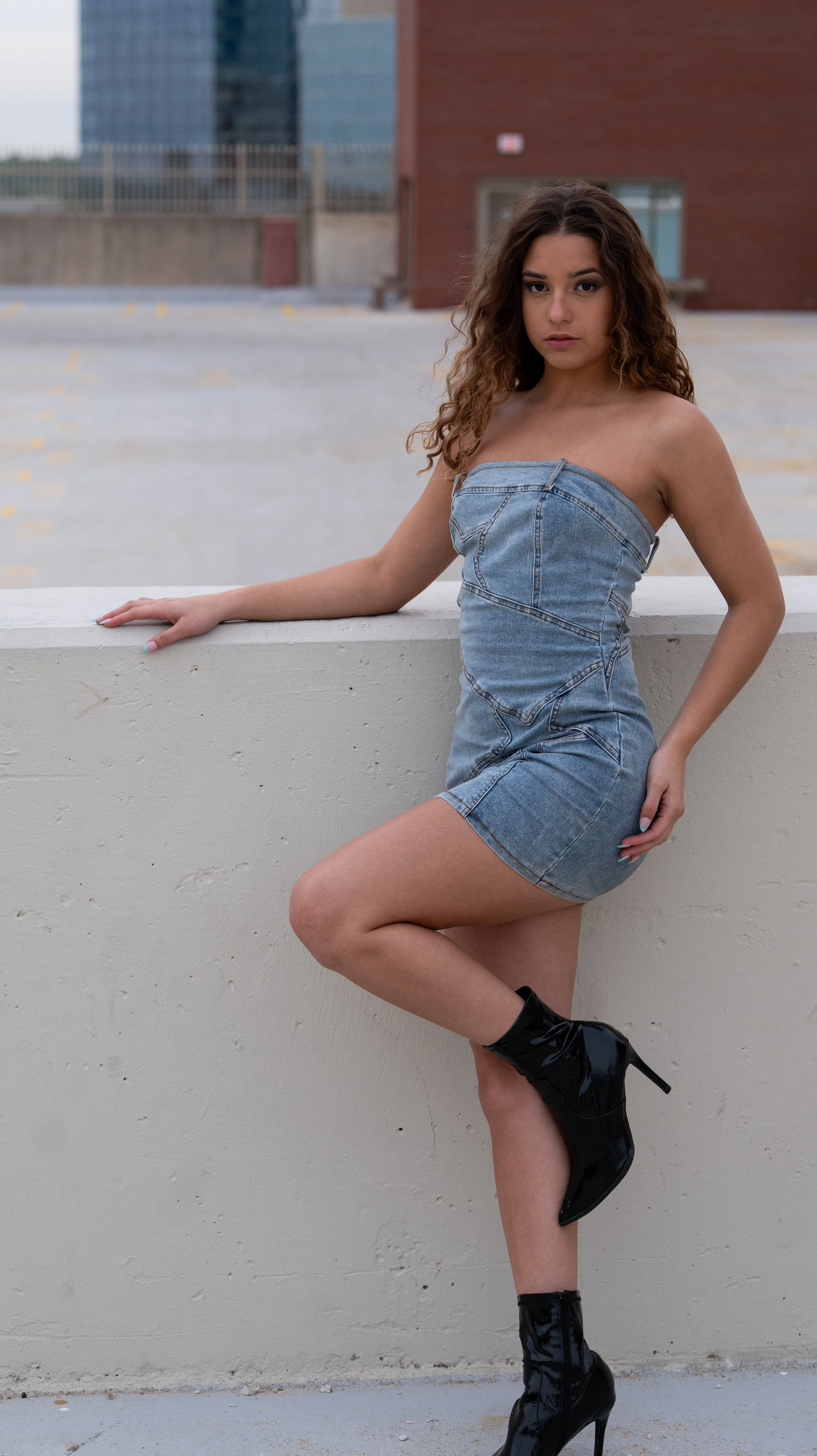 A young woman with curly brown hair and fair skin standing outdoors on a rooftop, wearing a strapless denim dress and black high-heeled boots, posing against a concrete railing with a cityscape background.