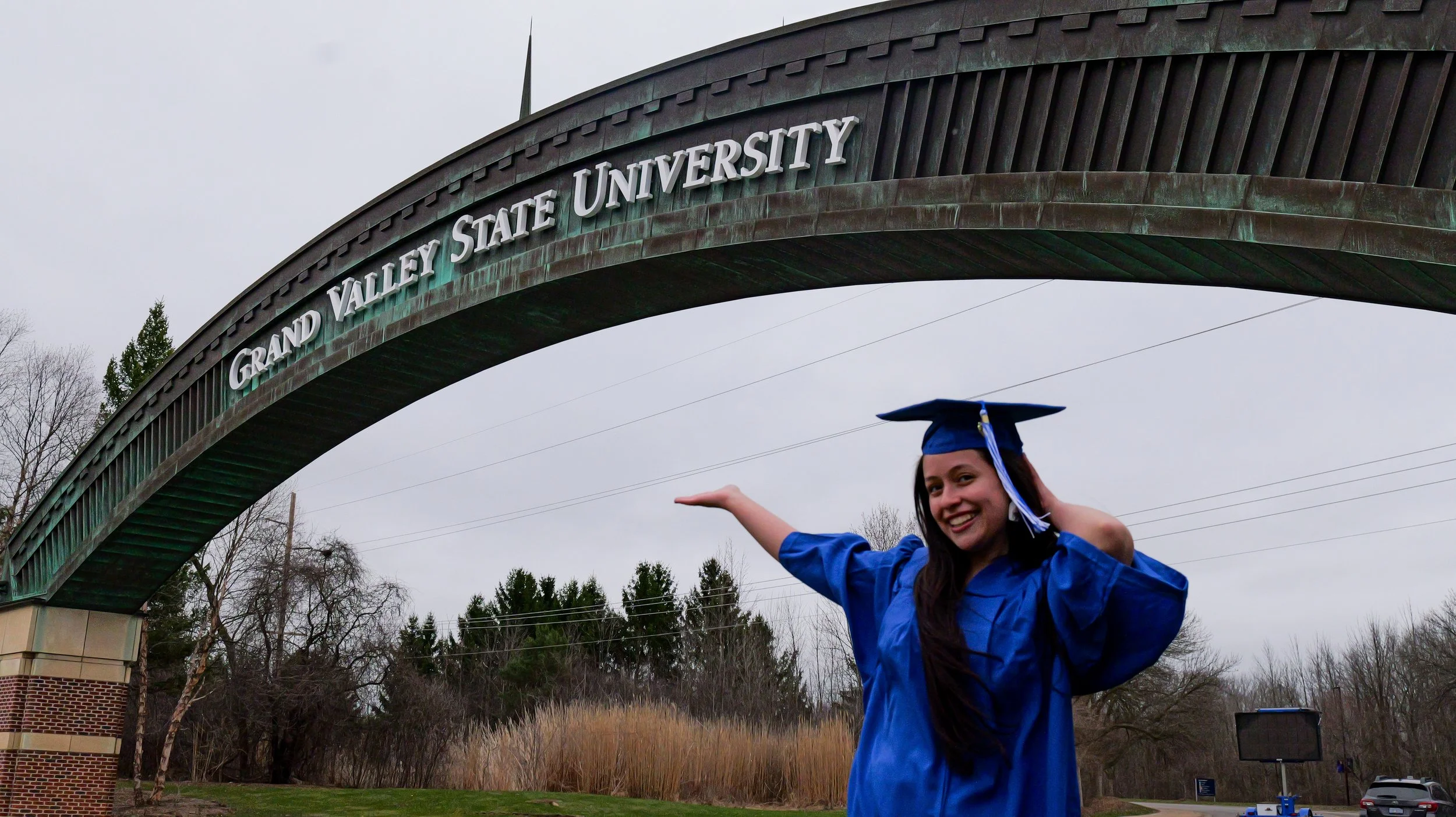 A young woman in blue graduation gown and cap smiling and gesturing towards a sign that reads 'Grand Valley State University' on an archway outside the university campus.