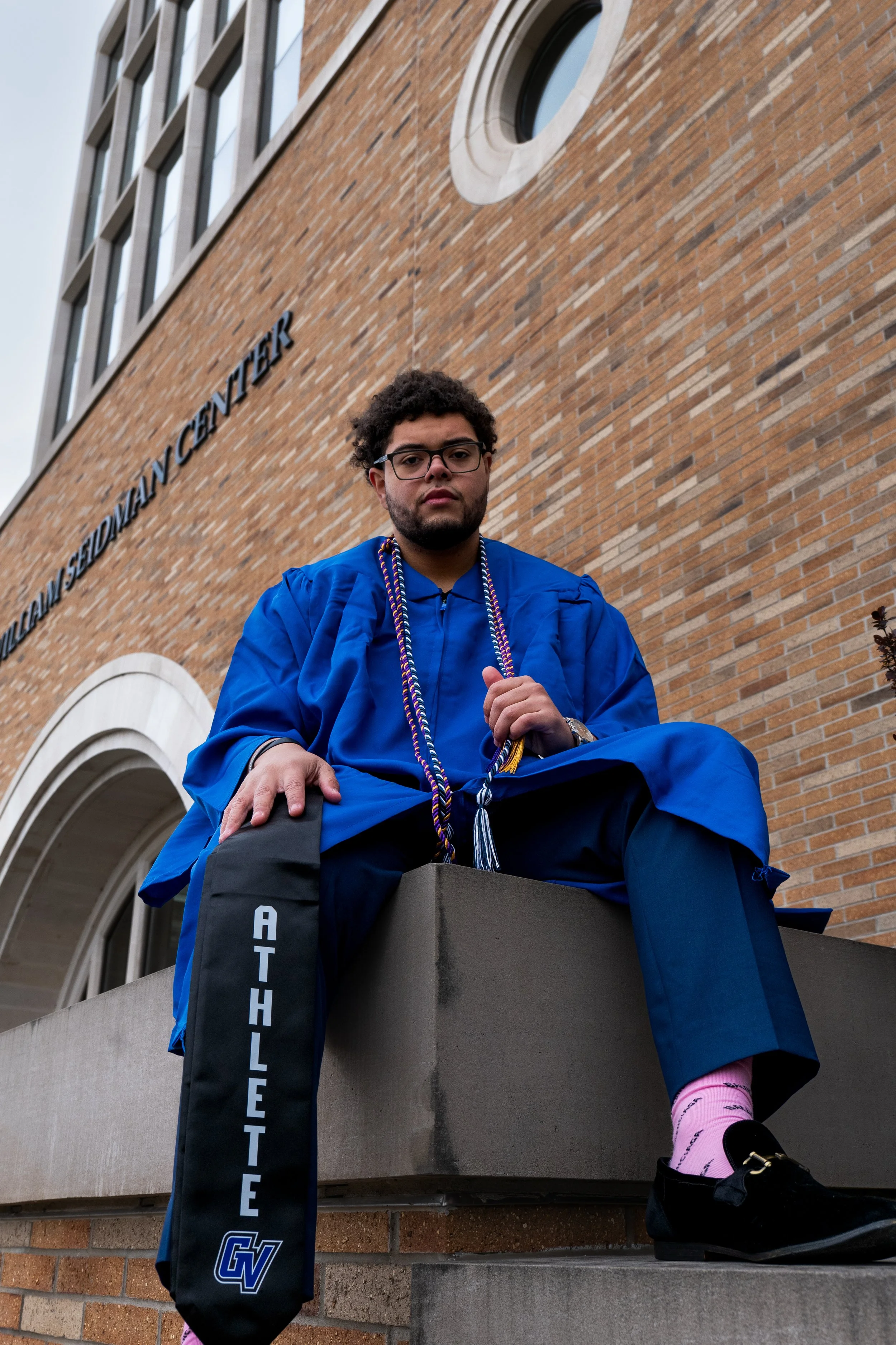Young man in blue graduation gown and cap sitting on a concrete ledge outside a brick building, with a hanging tassel and glasses, and a bag with "GW" logo hanging from his arm.