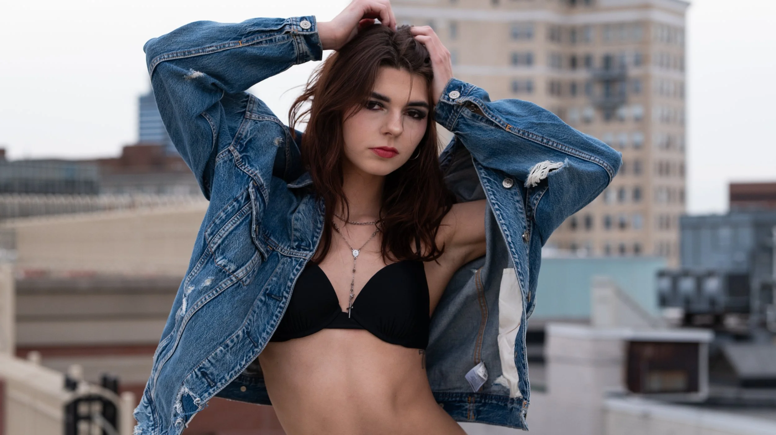 A young woman with long submerged in a black bikini top, wearing a ripped denim jacket, standing on a rooftop in an urban setting with buildings in the background.