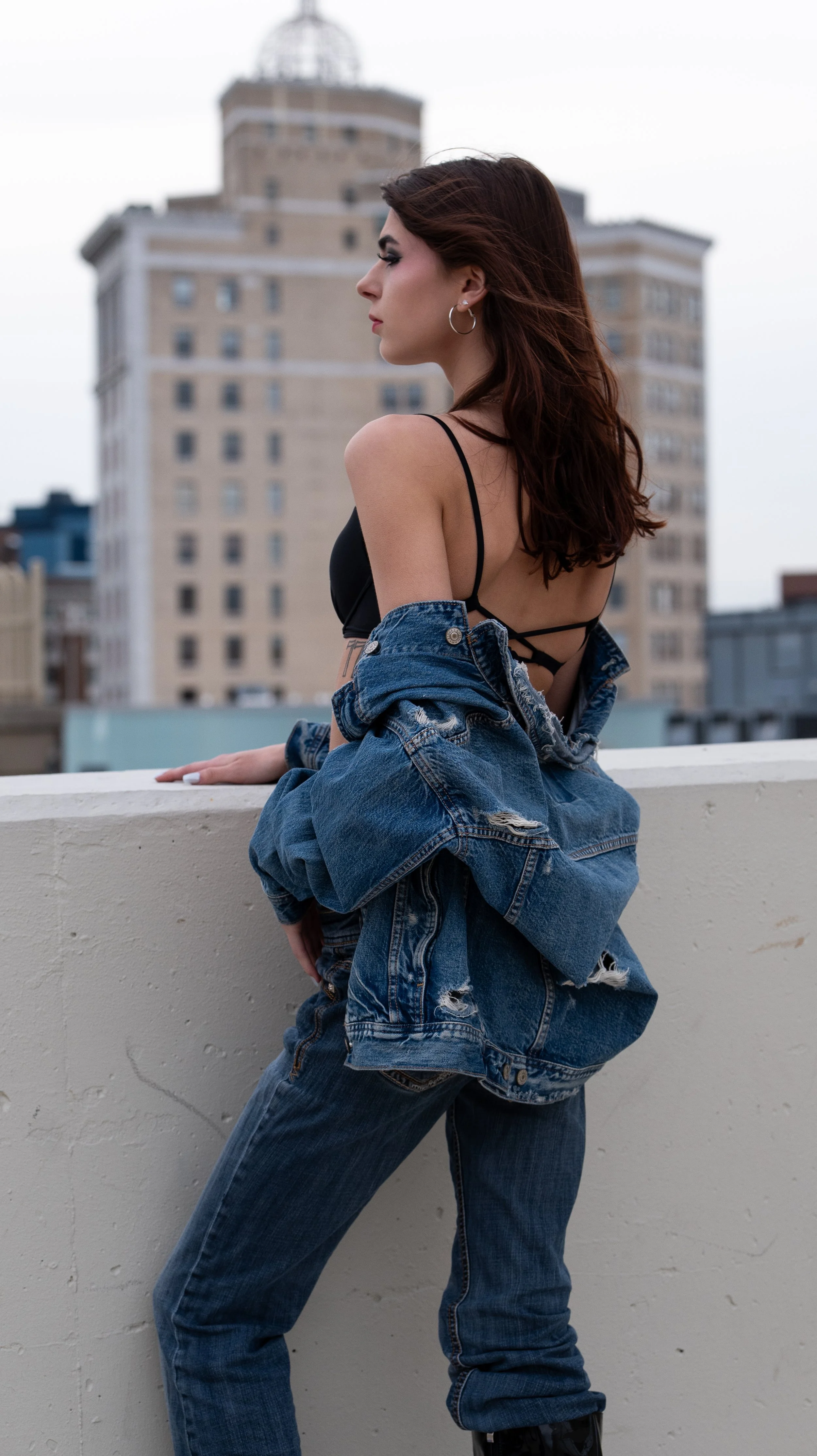 A woman with brown hair and hoop earrings, wearing a black top and over-sized denim jacket, standing on a rooftop with a cityscape in the background.