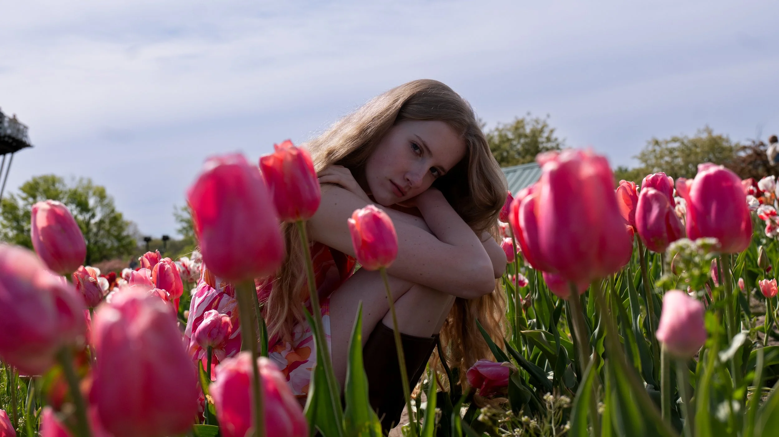 A young woman with long, wavy hair sitting in a field of pink tulips on a sunny day.