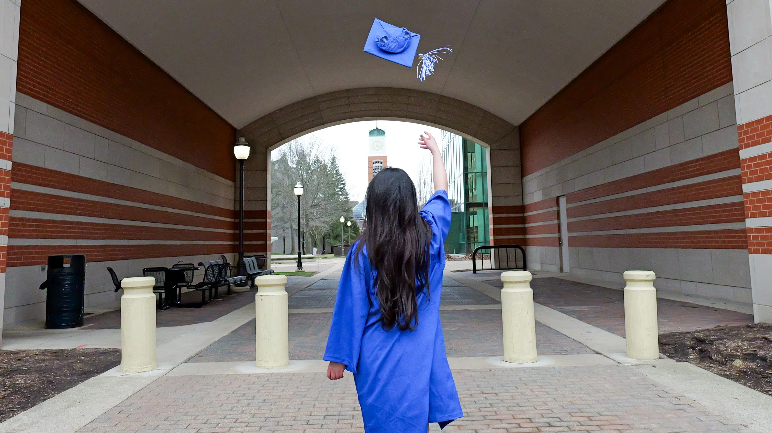 A woman in a blue graduation gown throws her cap in the air under a brick archway on a college campus, with a clock tower visible in the background.