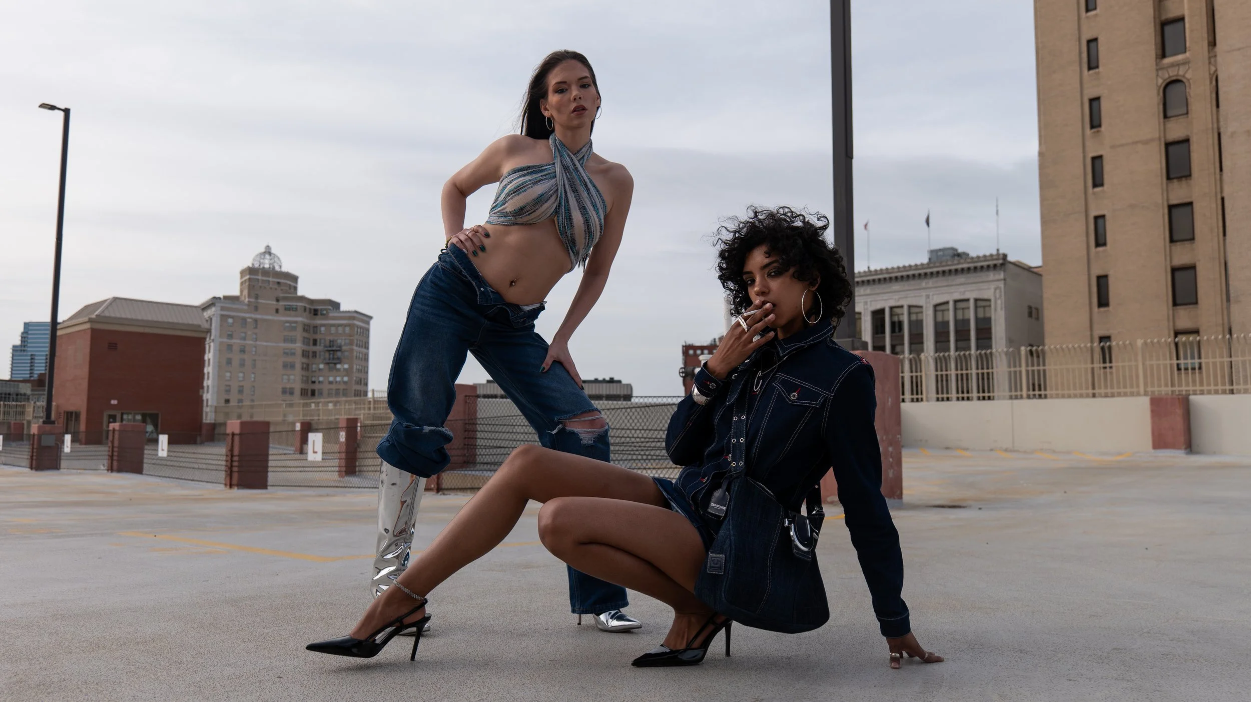 Two women are posing on a rooftop parking lot in an urban area with buildings and cloudy sky in the background. One woman is standing with one foot on a high heel, wearing a patterned halter top and ripped jeans. The other woman is sitting on the gro