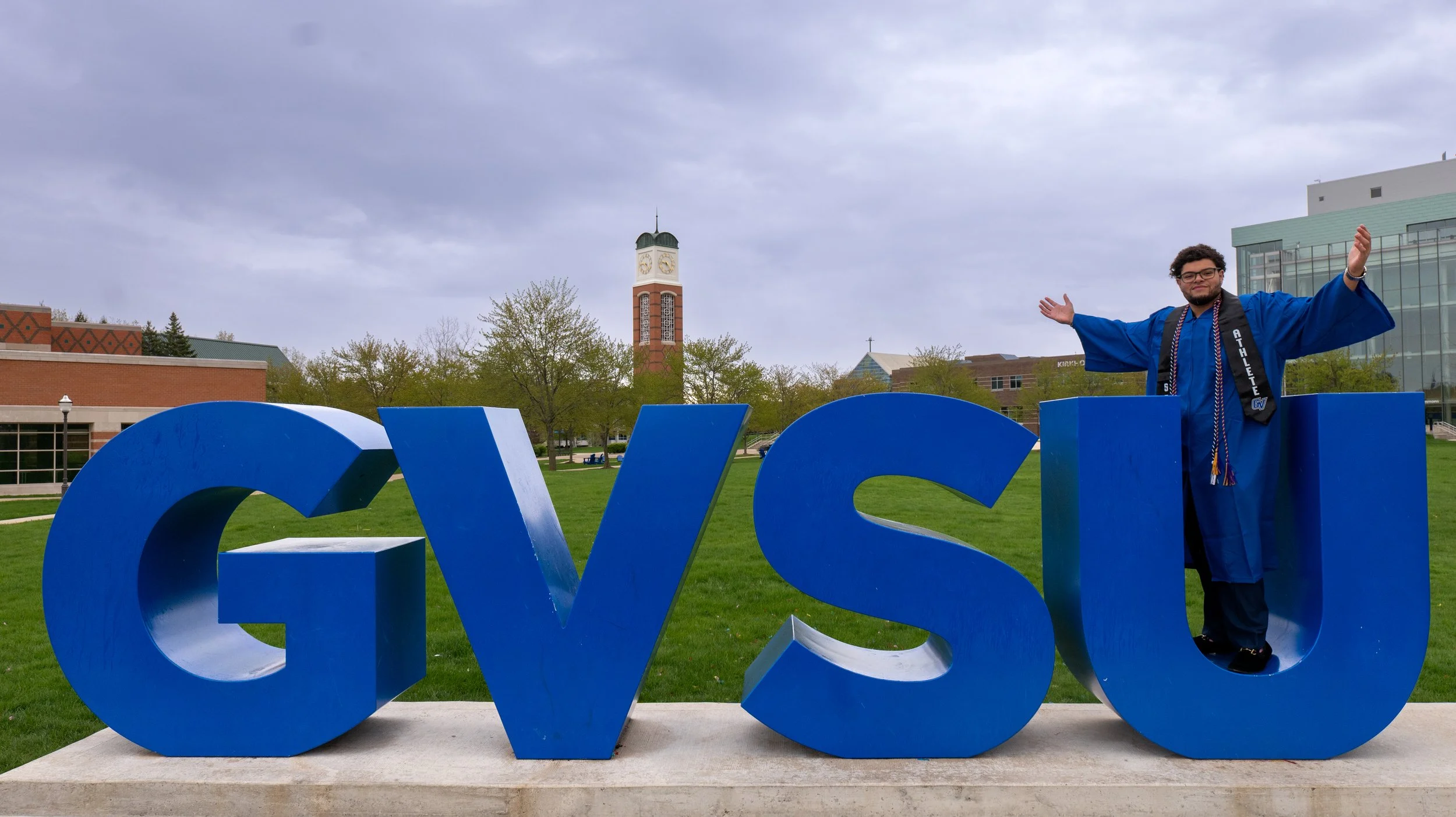 A person in a graduation gown standing behind large blue letters spelling 'CVS' on a grassy area with trees, a clock tower, and buildings in the background.