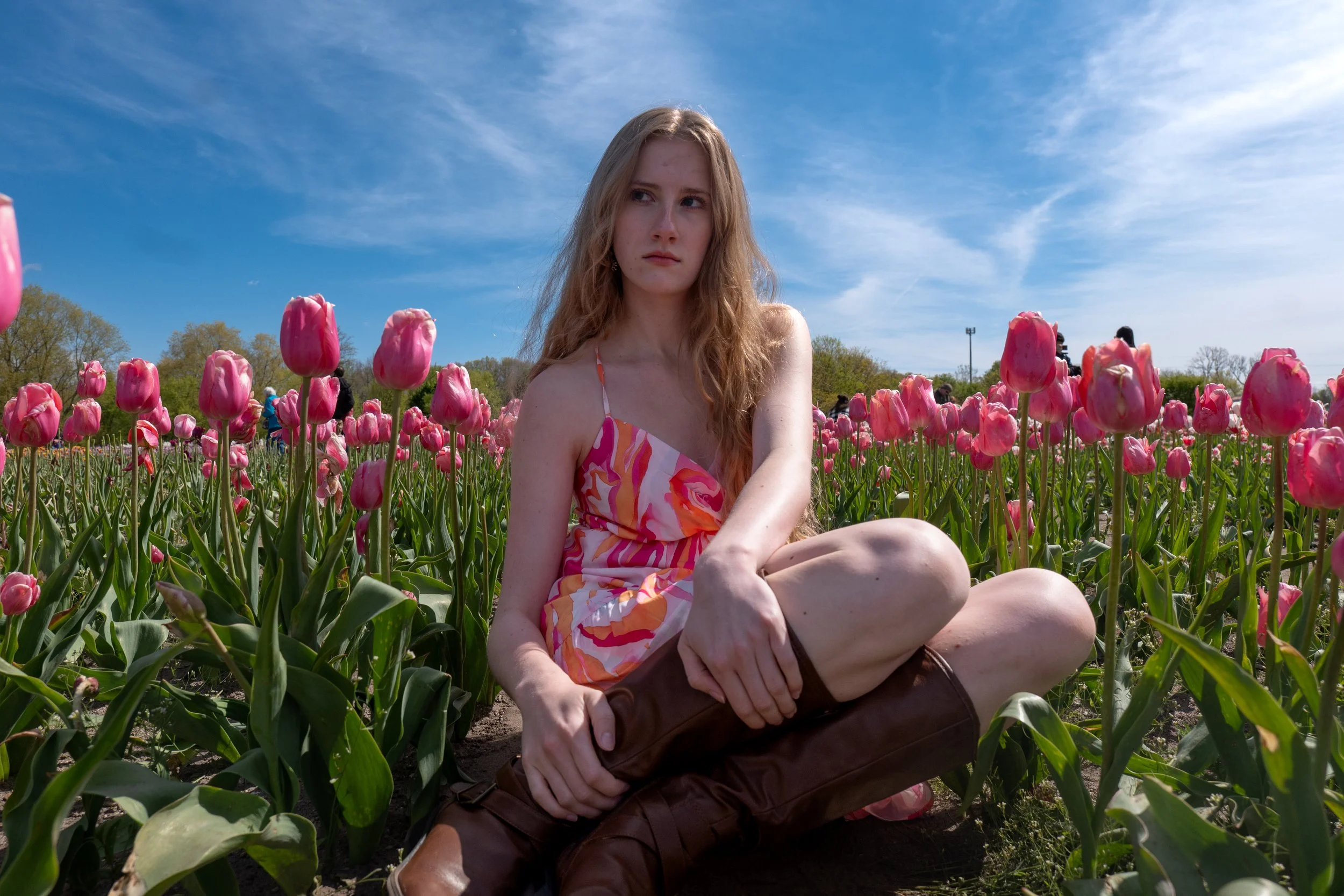 A young woman with long curly hair sitting in a field of pink tulips under a bright blue sky with some wispy clouds.