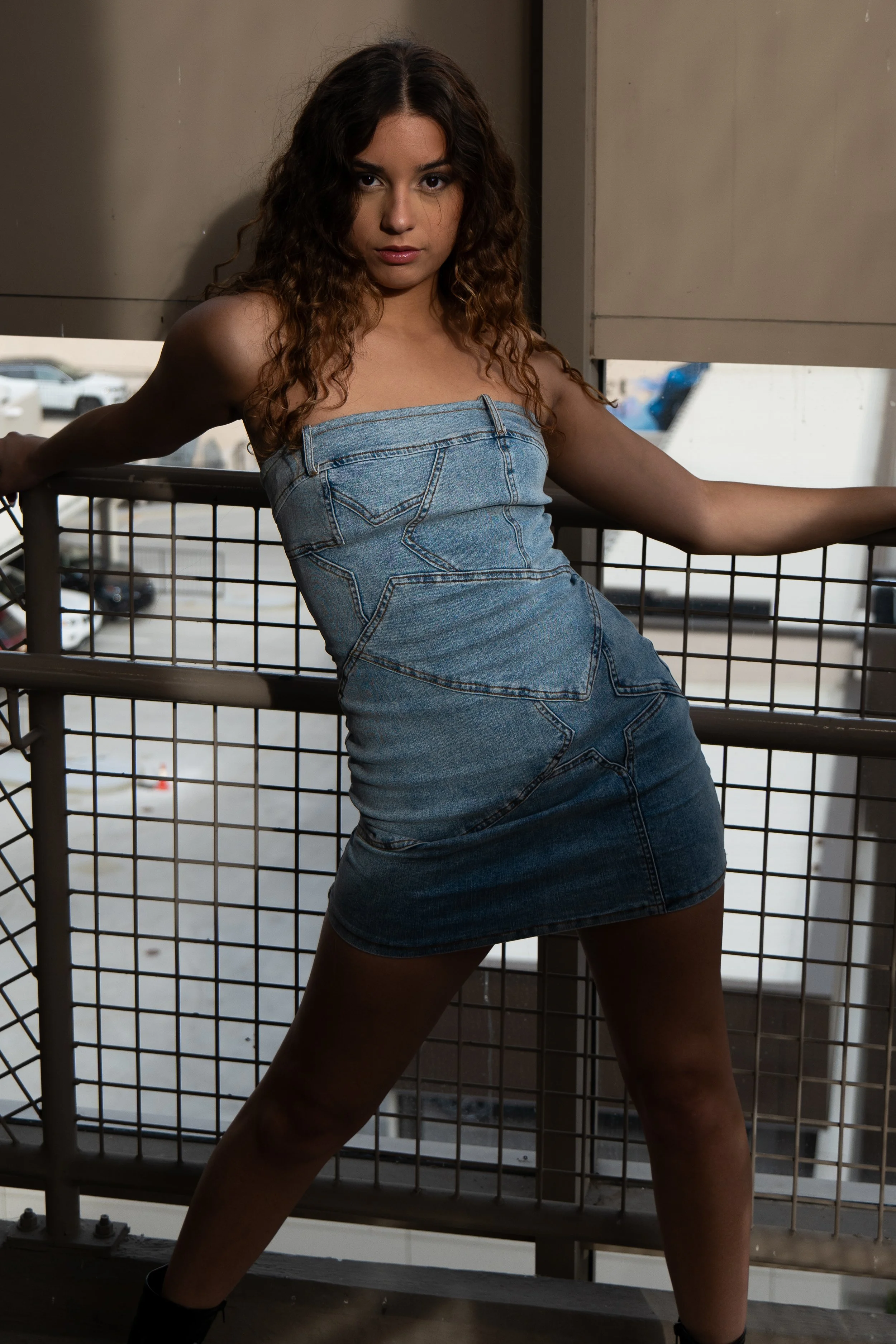 Young woman with long curly hair posing in front of a metal railing on a balcony, wearing a denim dress.