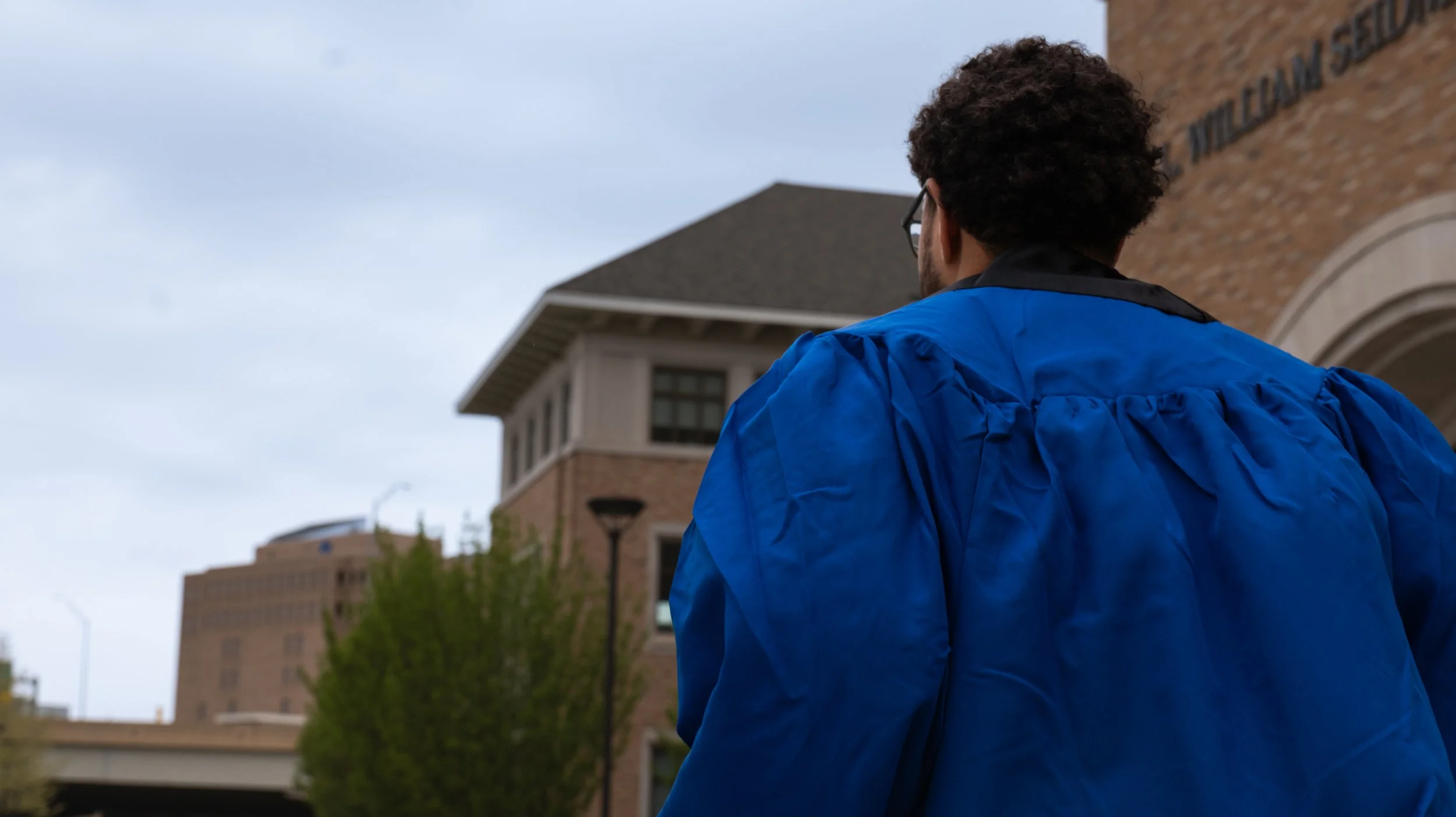 Back view of a man with curly hair wearing glasses and a blue jacket, standing outdoors near a brick building with partially cloudy sky.