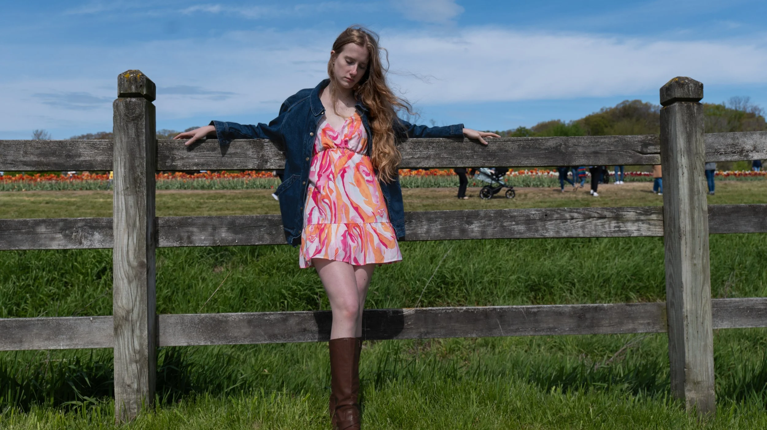A young woman with long, wavy red hair wearing a colorful pink, orange, and white patterned dress, a denim jacket, and tall brown boots, standing outdoors near a wooden fence with her eyes closed and arms resting on the fence, in a field with green g