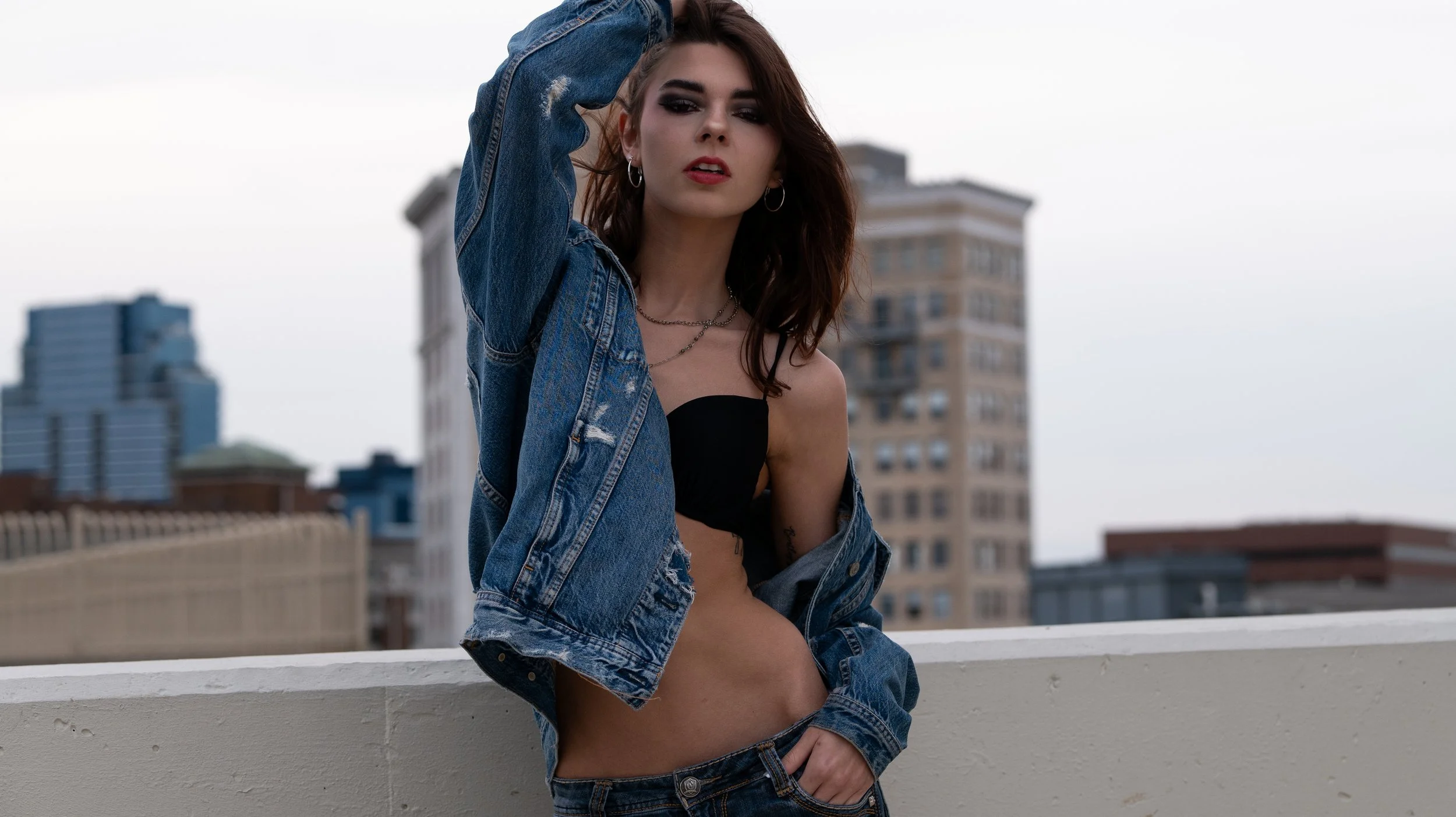A woman with red hair and makeup, wearing a black crop top and jeans with a denim jacket, sitting on a rooftop with city buildings in the background.
