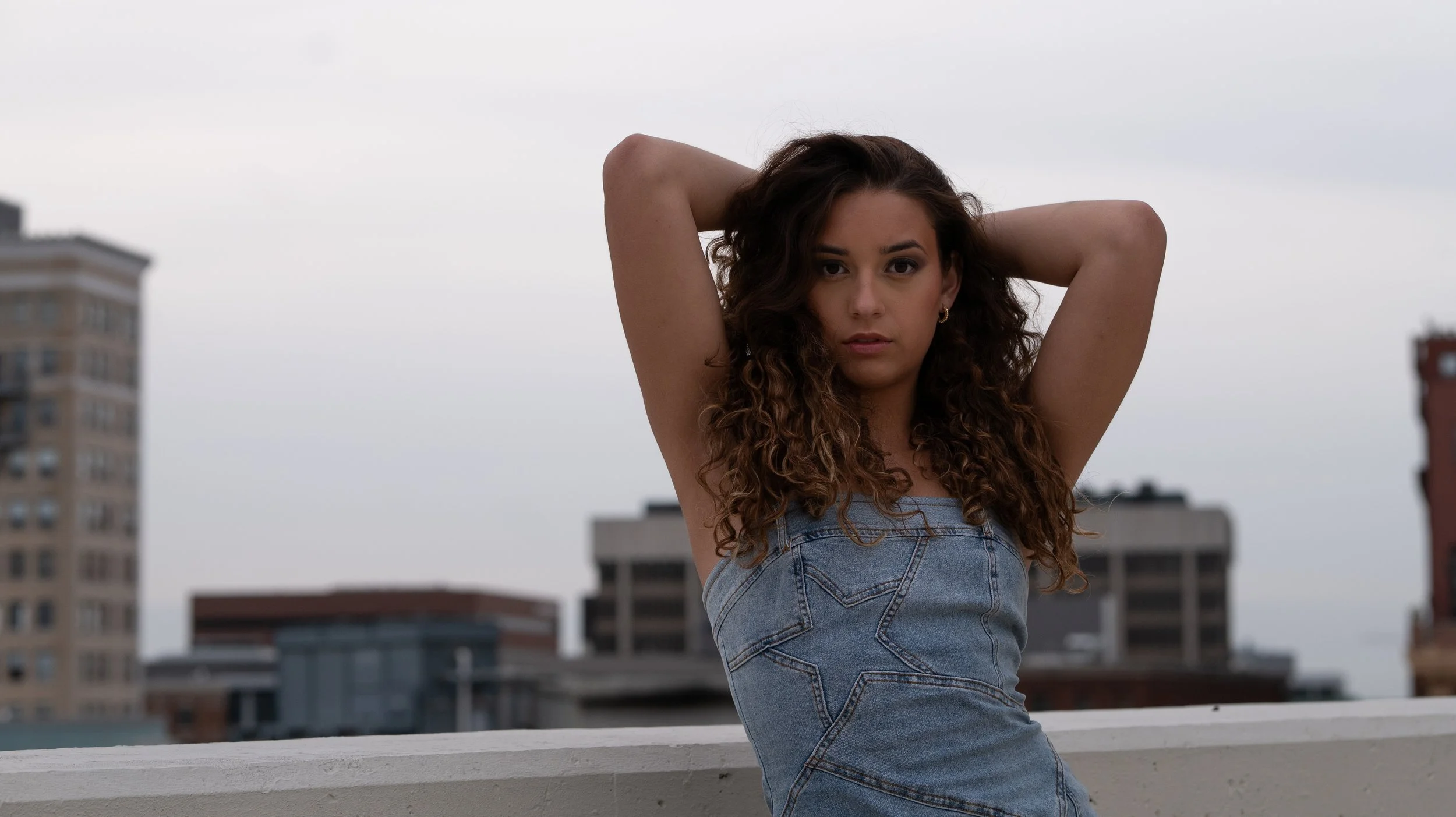 A young woman with curly brown hair and hoop earrings stands on a rooftop with her arms raised behind her head, wearing a denim tank top, with a cityscape of tall buildings in the background on a cloudy day.