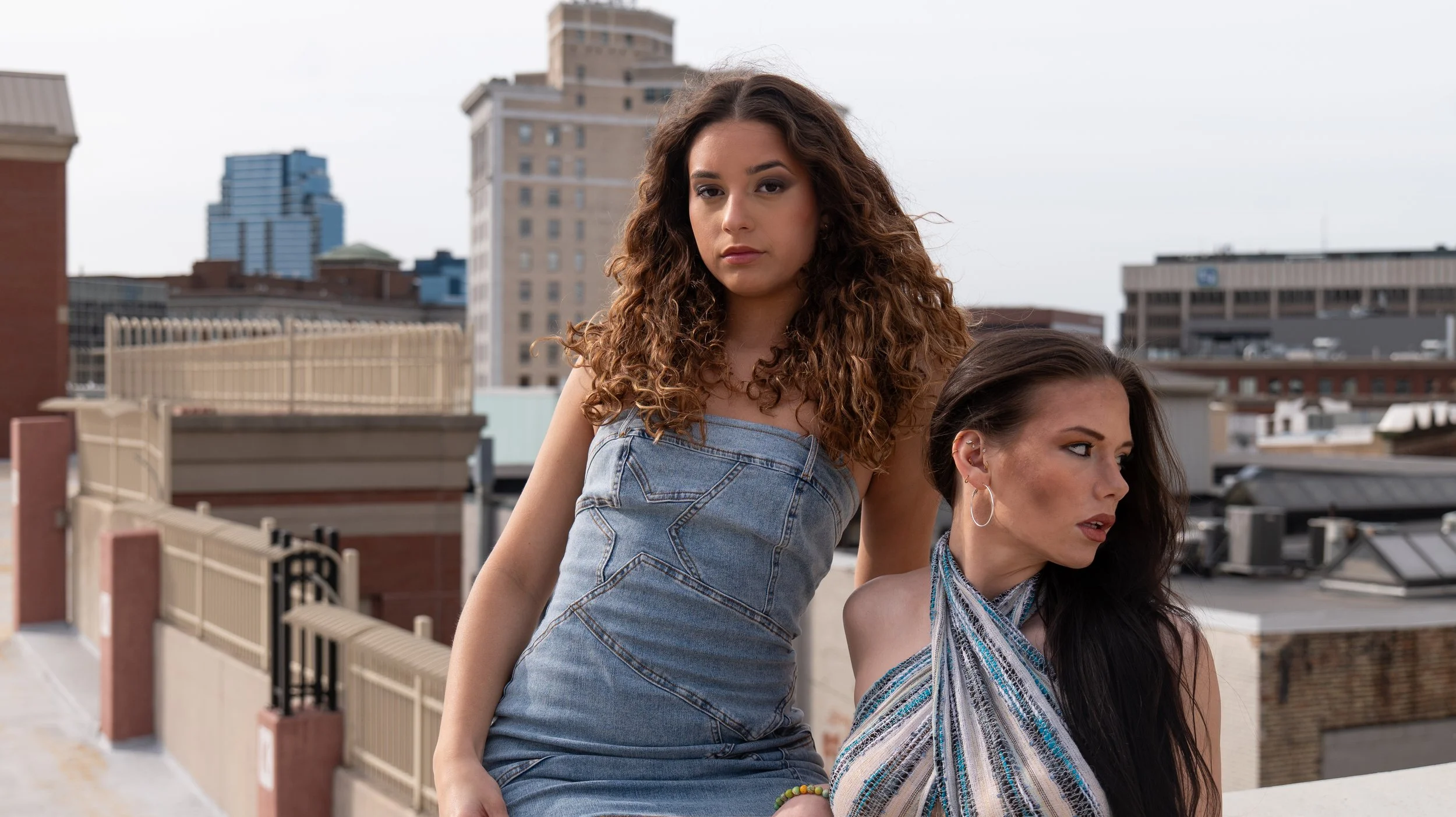 Two women with long hair on a rooftop with city buildings in the background.