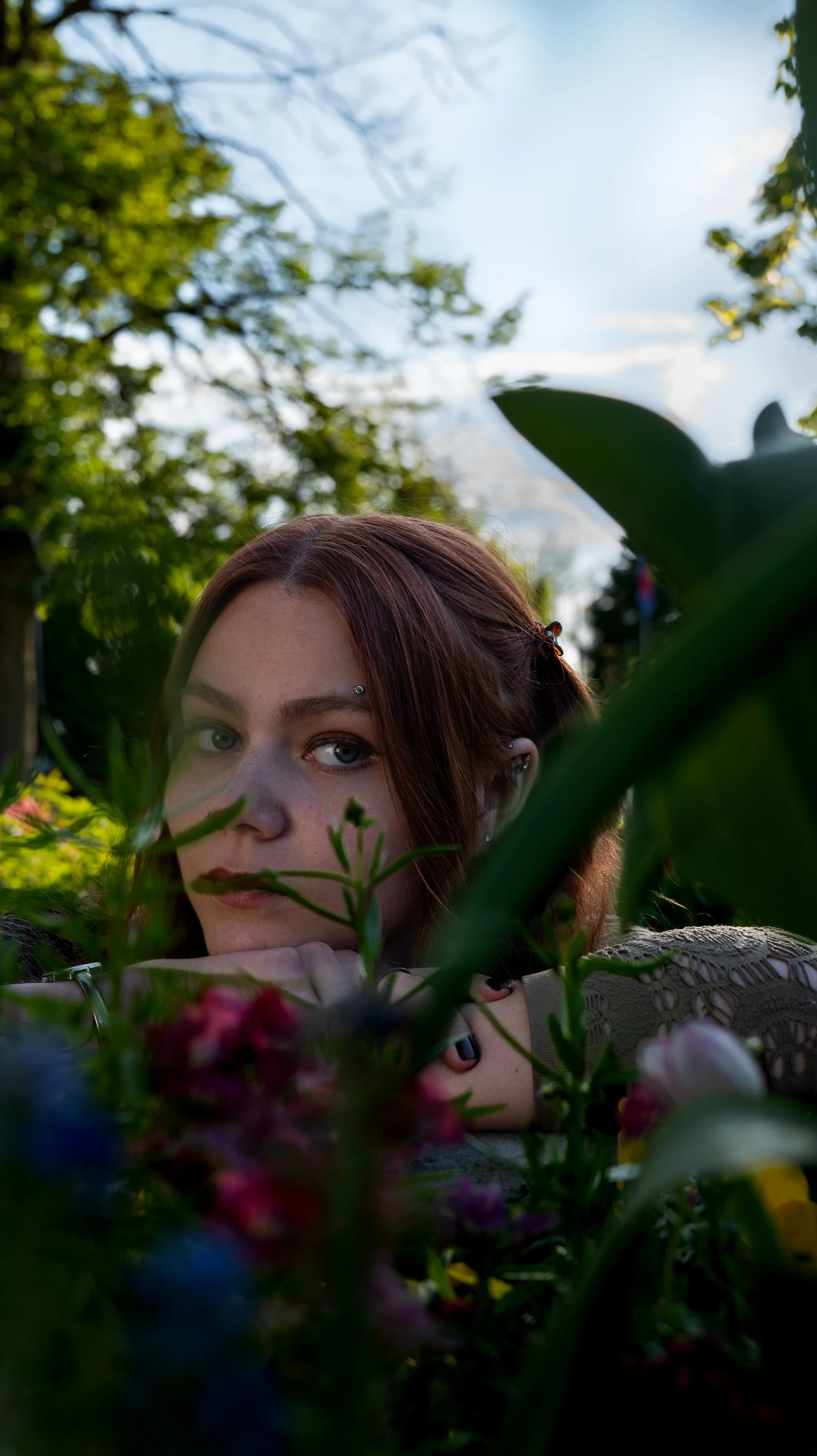 A young woman with reddish-brown hair and blue eyes lying on her arms behind lush greenery and flowers in a garden. She is looking directly at the camera with a contemplative expression.