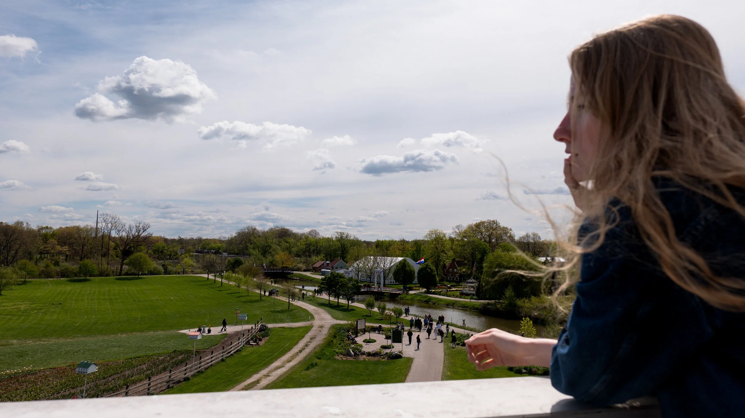 A woman with long hair looking out over a park with trees, a pond, and buildings under a cloudy sky.