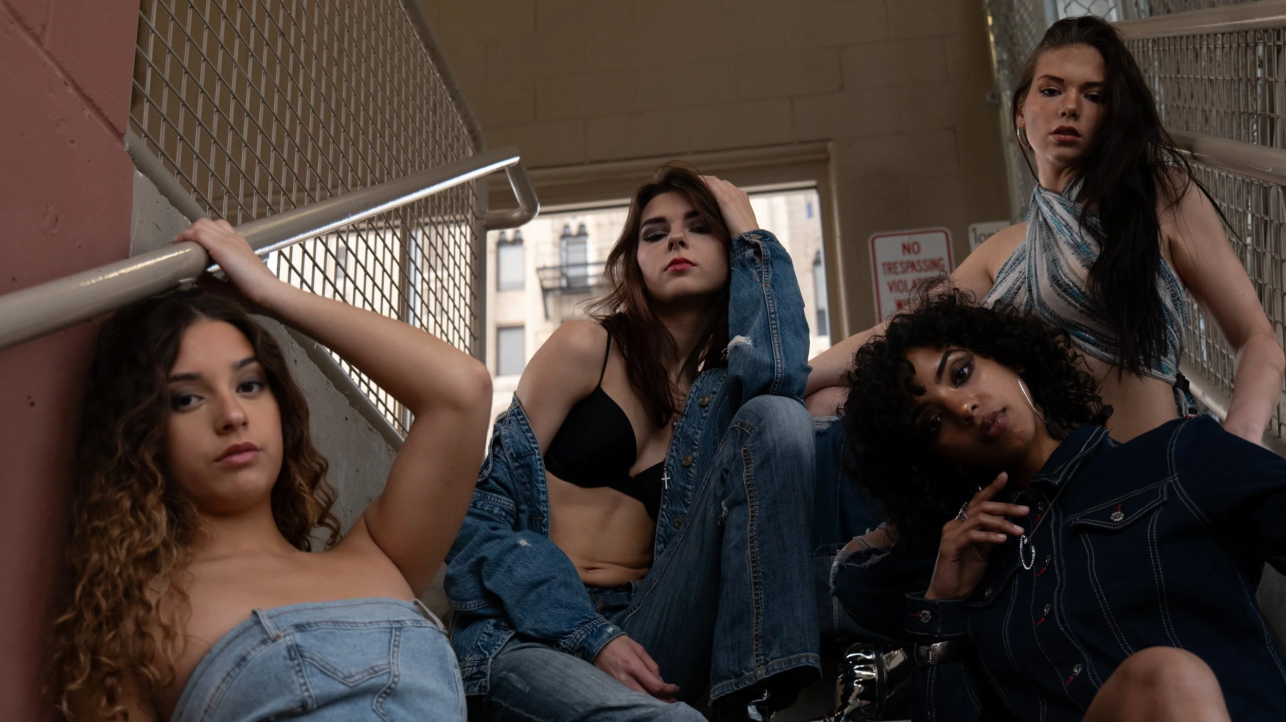 Four young women sitting on outdoor stairs at an urban location, posing with serious expressions. One has curly hair, another has straight dark hair, another has long dark hair, and the fourth has a short curly hairstyle. They are wearing casual clot