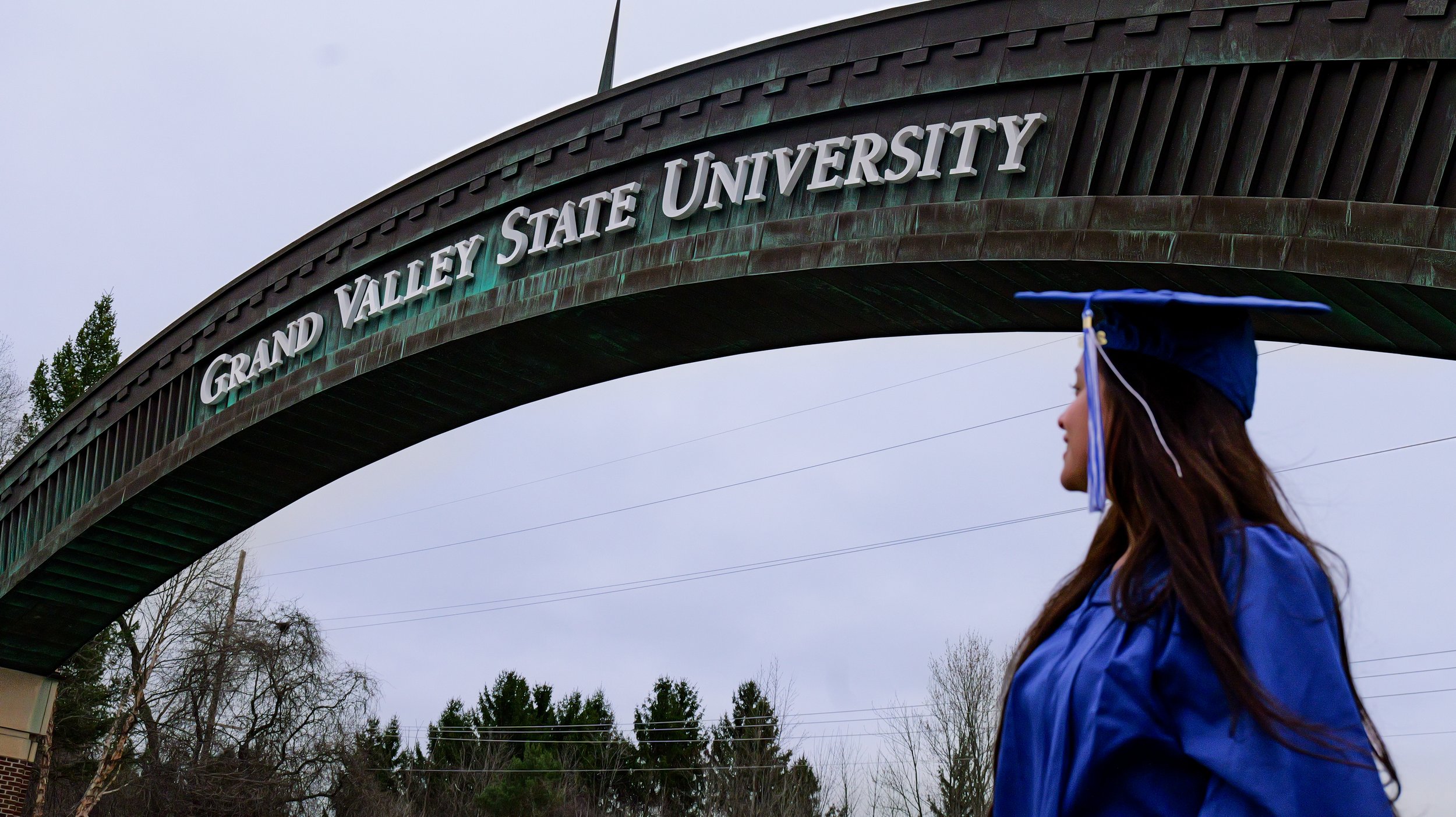 Student in blue cap and gown walking past Grand Valley State University arch on campus on a cloudy day.