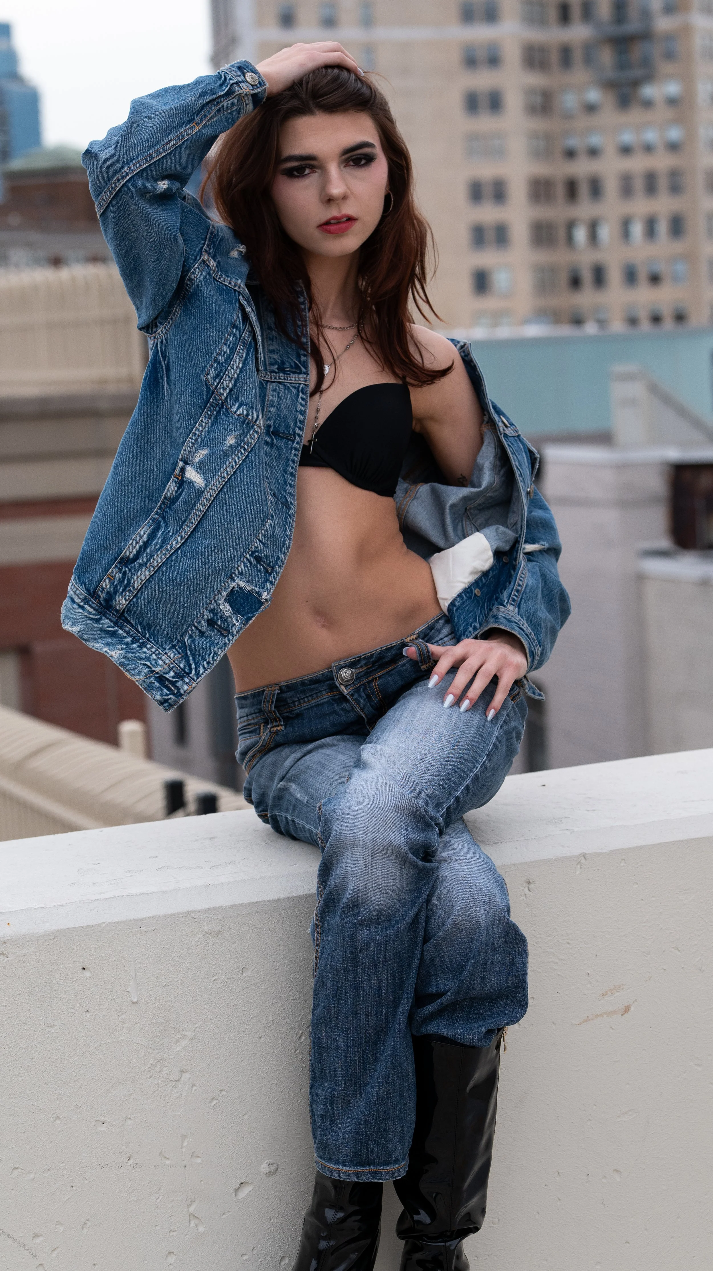 Young woman with auburn hair posing on a rooftop, wearing a denim jacket, black bra, ripped jeans, and black boots, with city buildings in the background.