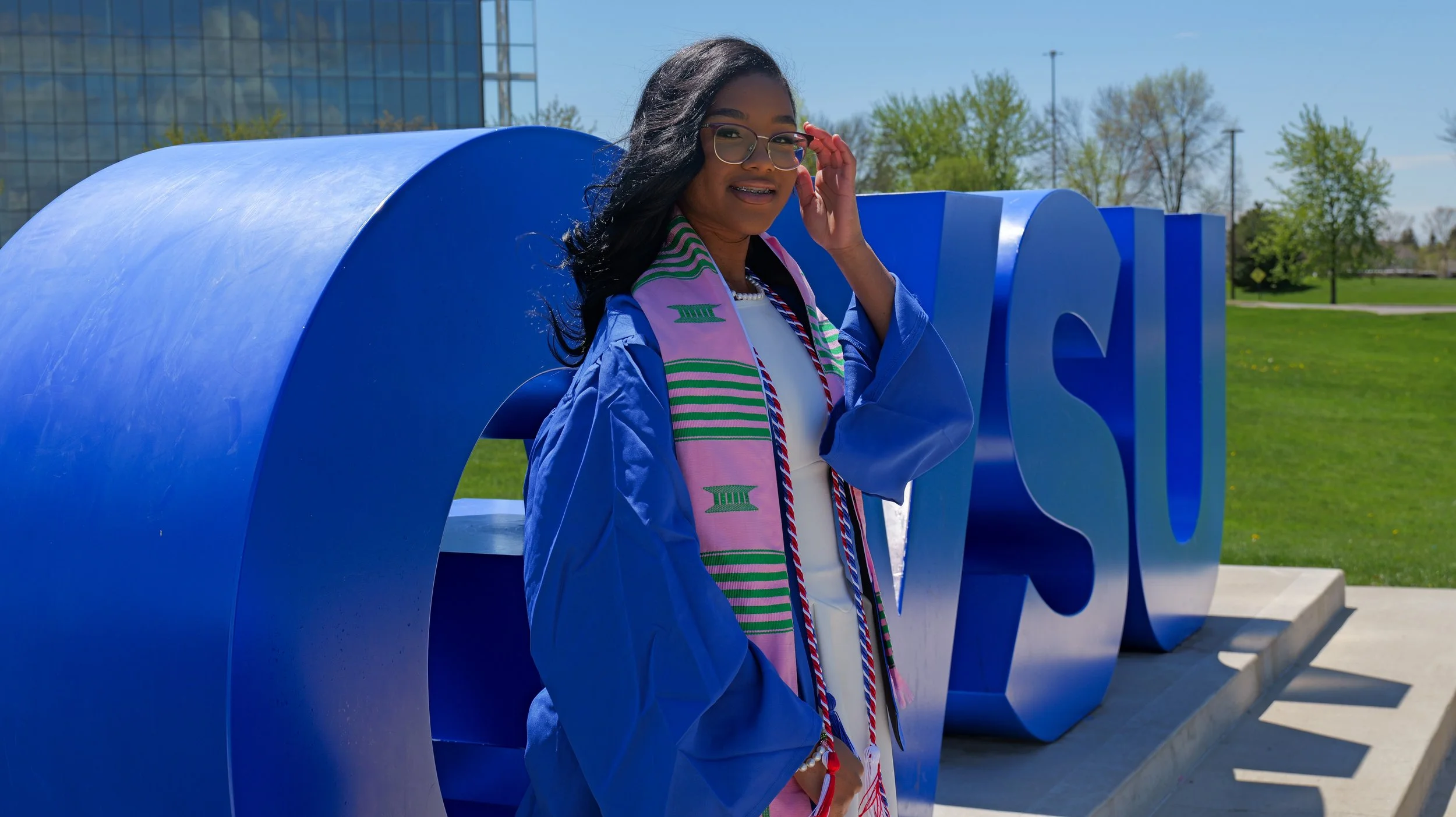 Young woman in a graduation gown and stole standing outdoors next to a large blue sculpture, smiling and adjusting her glasses on a sunny day.
