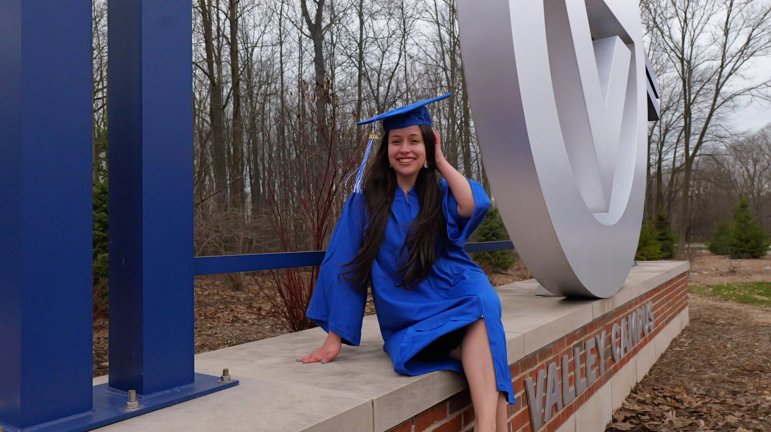 A young woman in a blue graduation gown and cap sitting on a brick ledge next to a large white emblem, smiling and touching her head outdoors with leafless trees in the background.