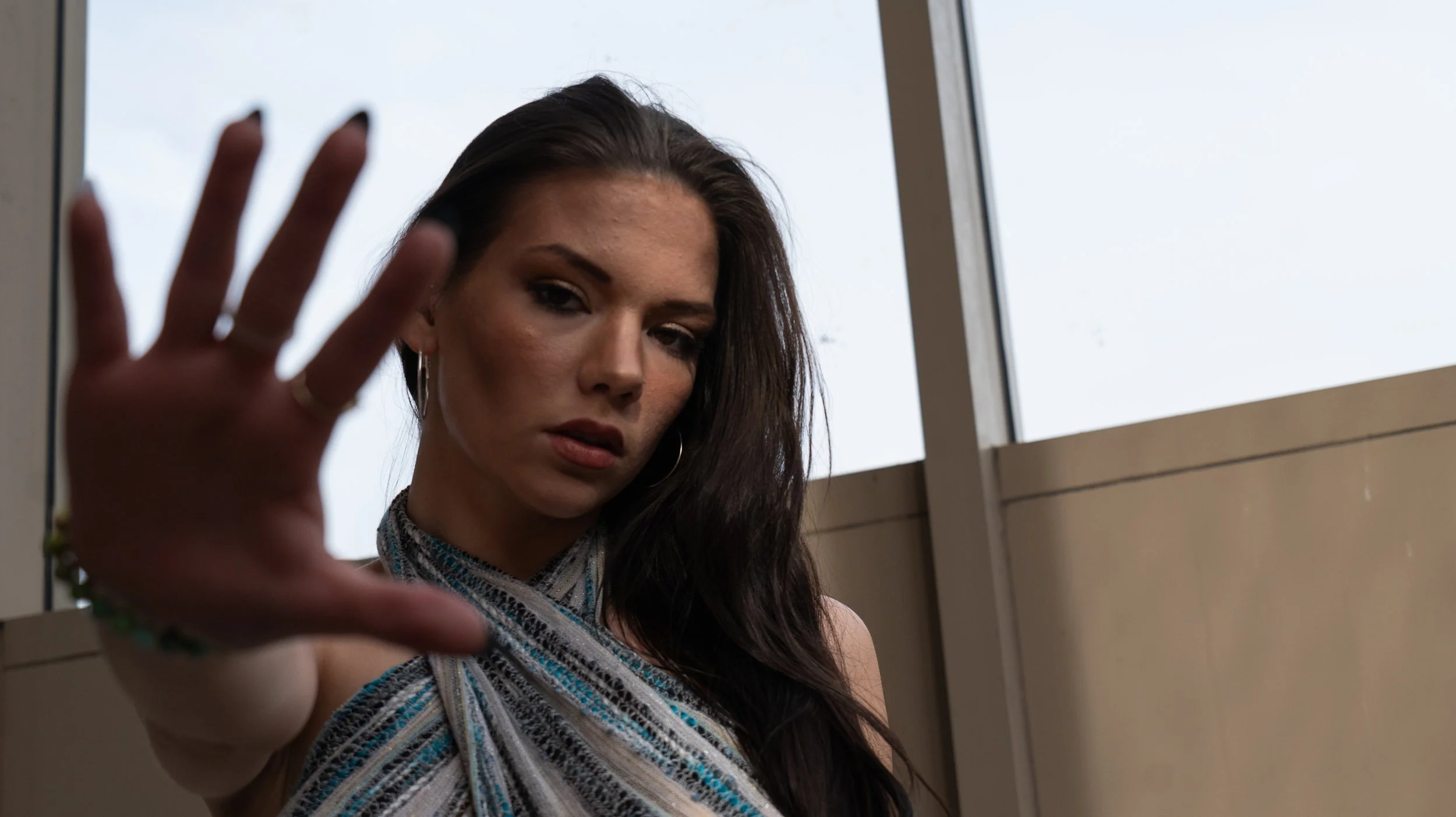 A woman with long dark hair, wearing hoop earrings and a colorful, striped top, leaning against a window and raising her hand towards the camera.