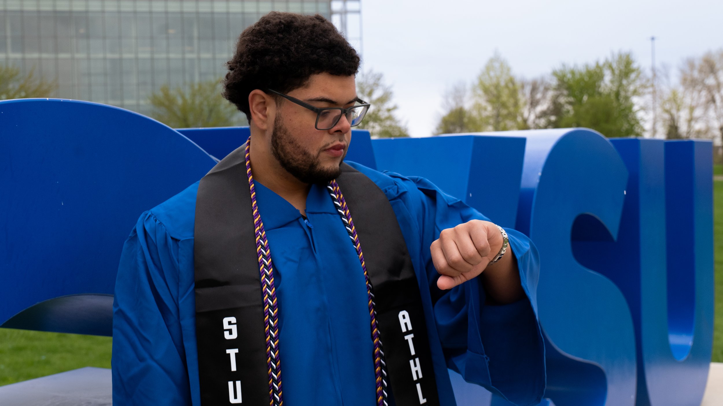 A young man in an academic gown and cap looking at his watch during a graduation ceremony outdoors.