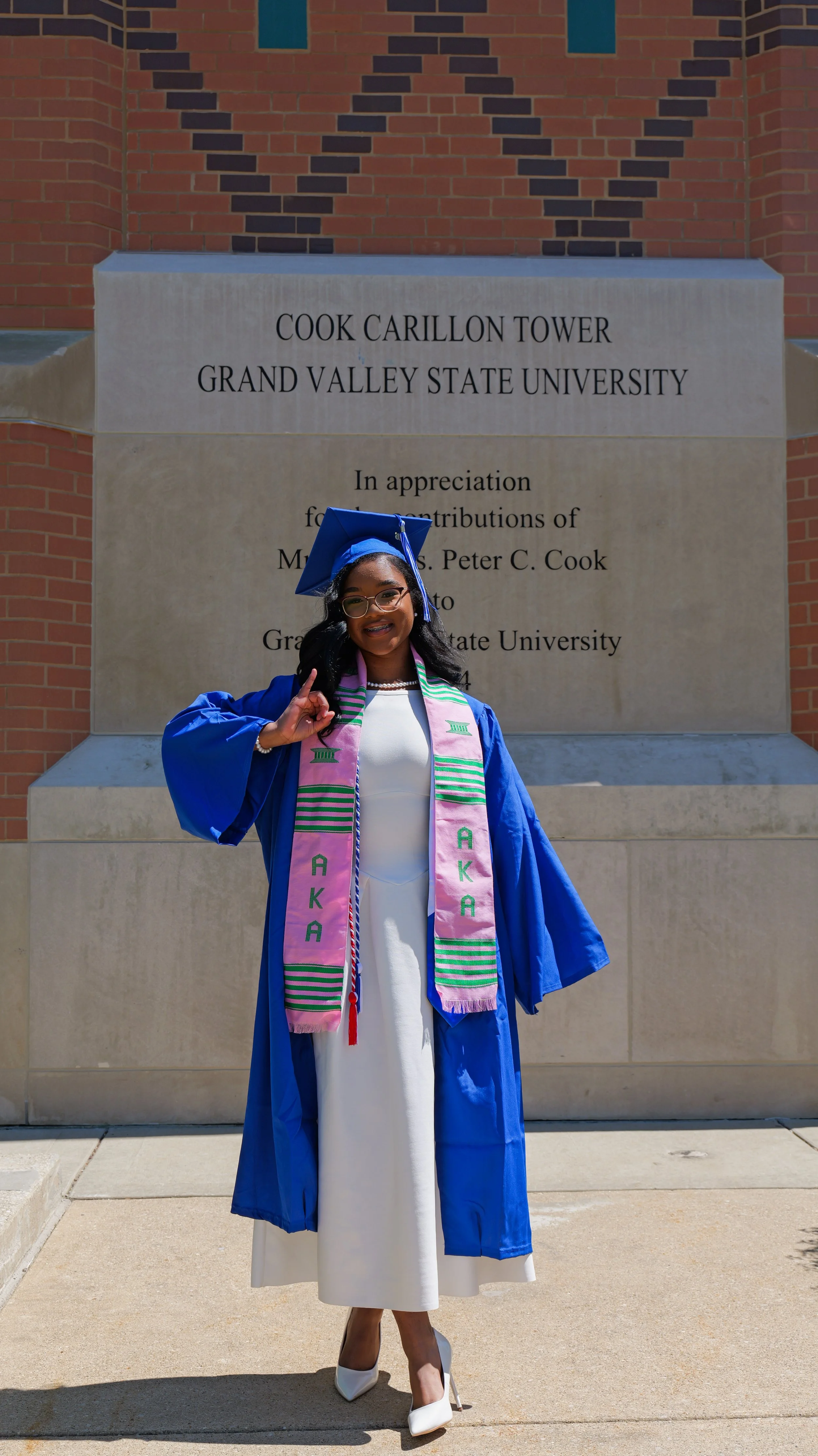 Female college graduate wearing a blue cap and gown, standing in front of a campus monument with a brick wall, smiling and making a gesture with her right hand.