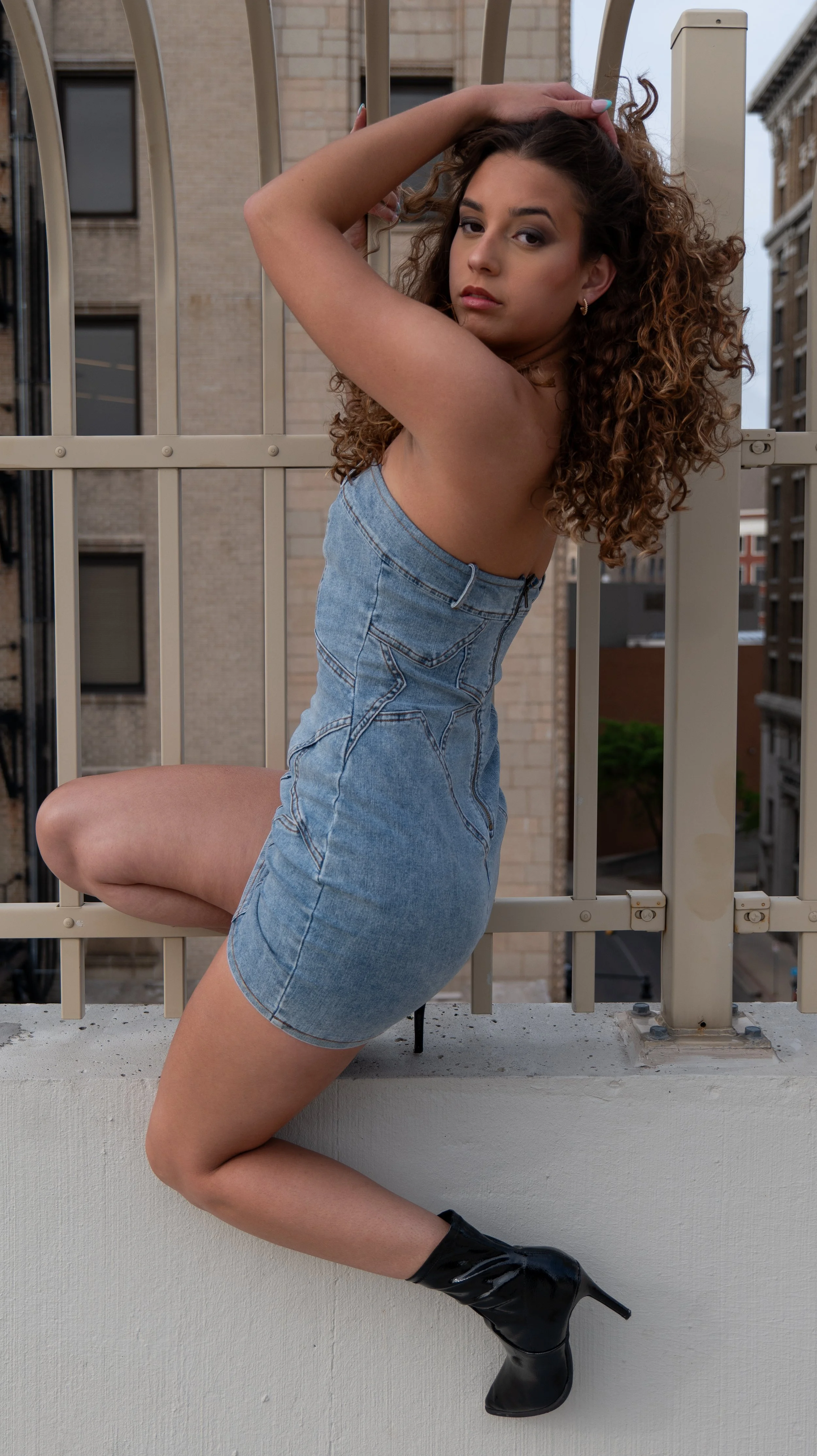 A woman with curly hair wearing a denim dress and black high-heeled boots poses on a balcony railing in an urban setting.