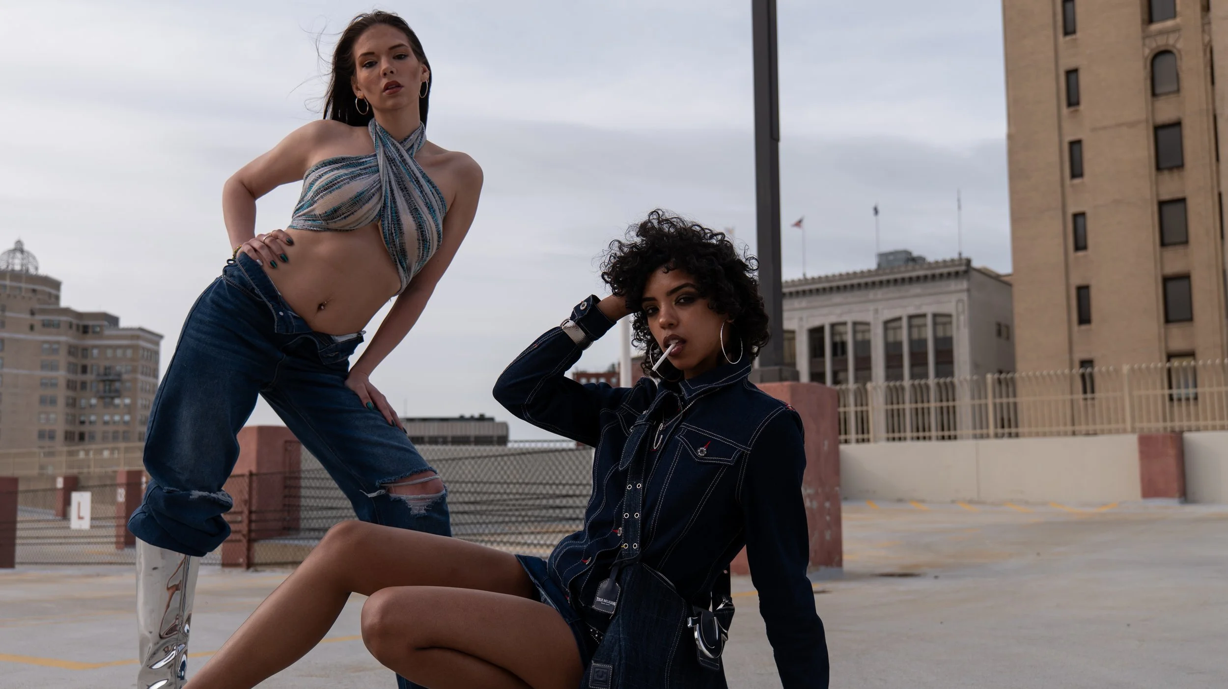 Two young women with dark hair and hoop earrings posing on a rooftop parking lot against a cityscape with buildings and cloudy sky; one woman standing with one hand on her hip, wearing ripped jeans and a striped halter top, and the other sitting, smo