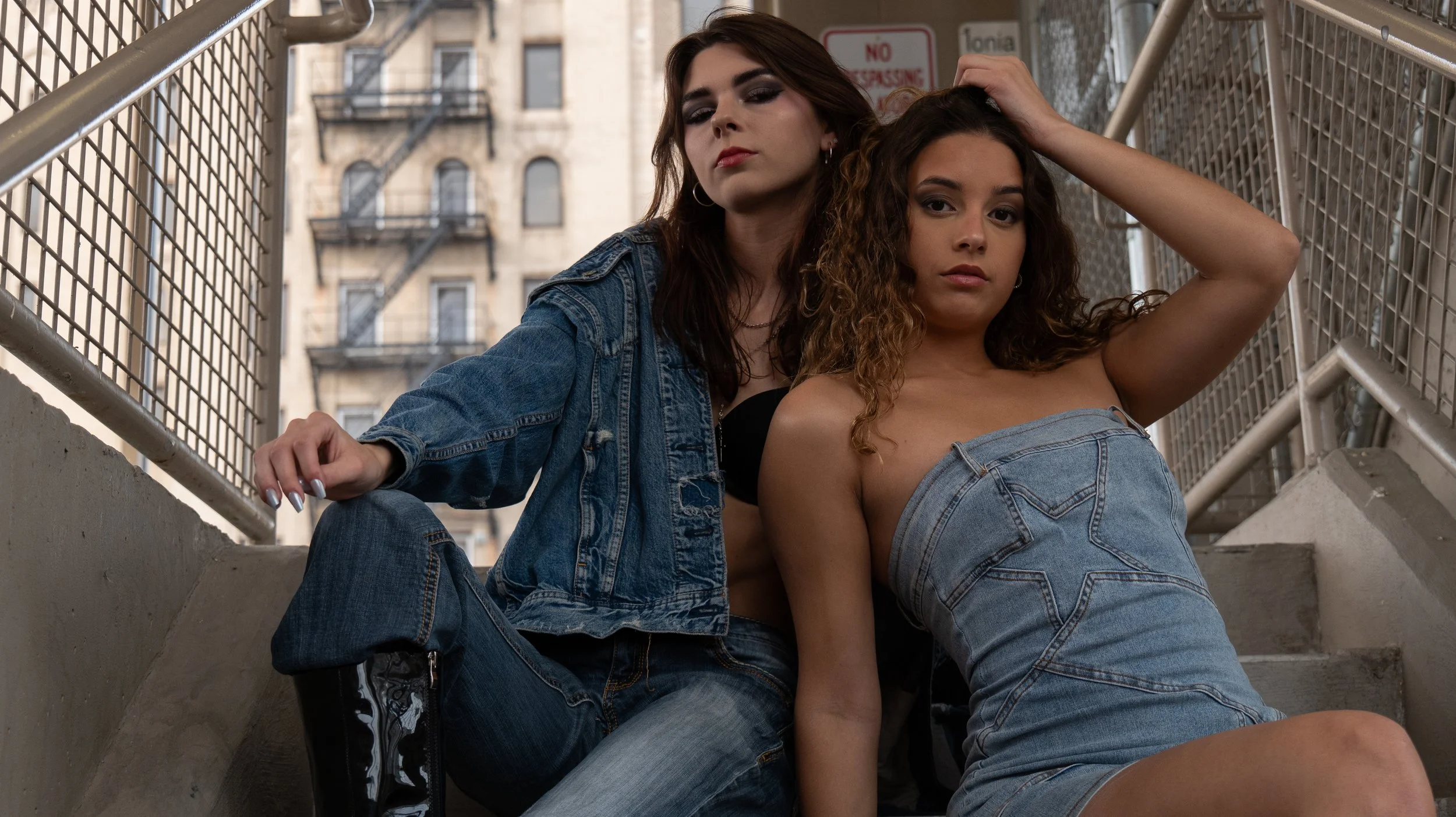 Two women sitting on outdoor stairs between metal railings with buildings in the background. One woman has long brown hair and the other has long curly hair. Both are wearing denim outfits.
