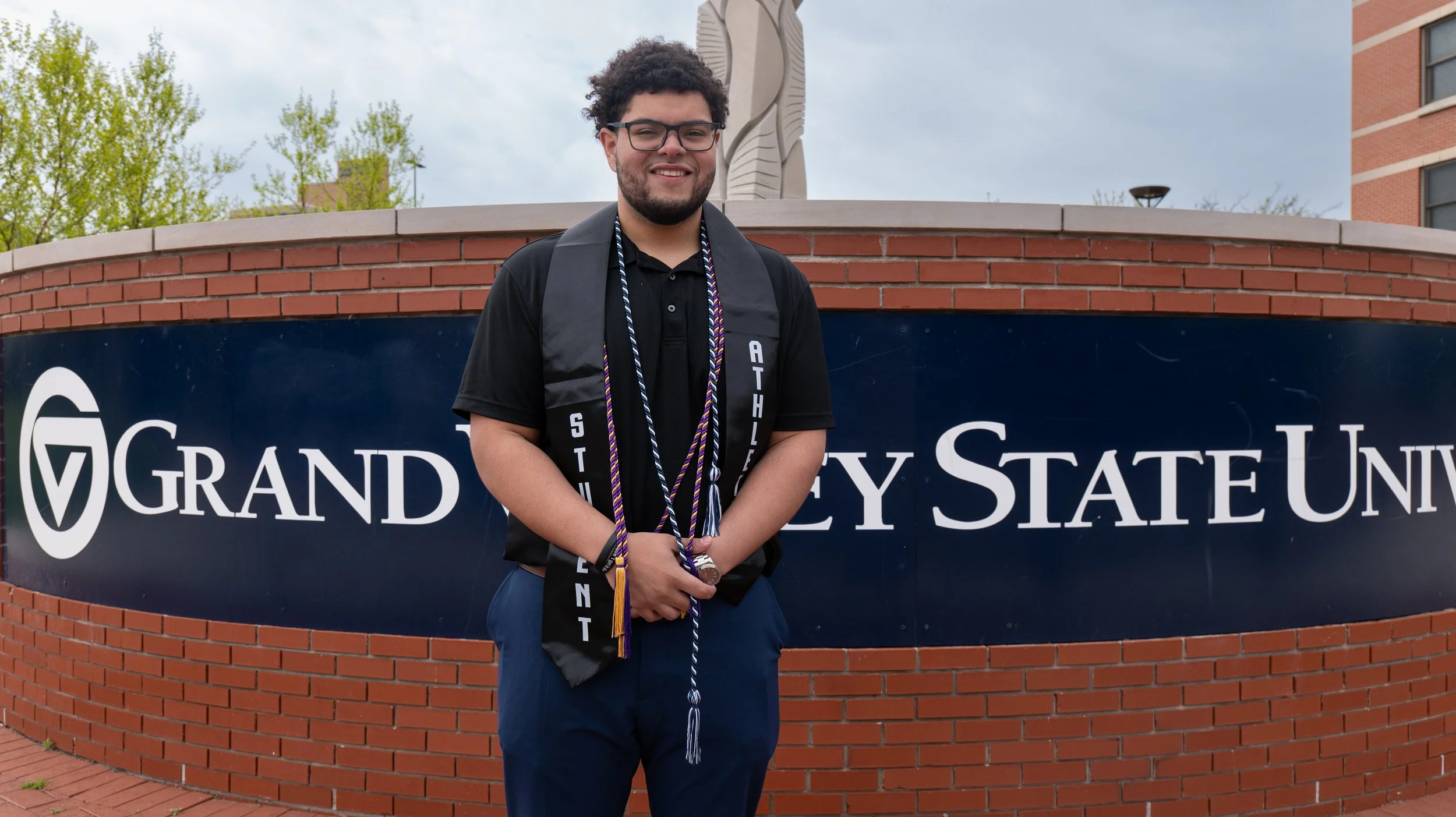 A smiling young man in glasses standing outdoors in front of a brick sign that reads 'Grand Valley State University' during daytime. He is wearing a black polo shirt, a black graduation sash with white text, and multiple cords around his neck.