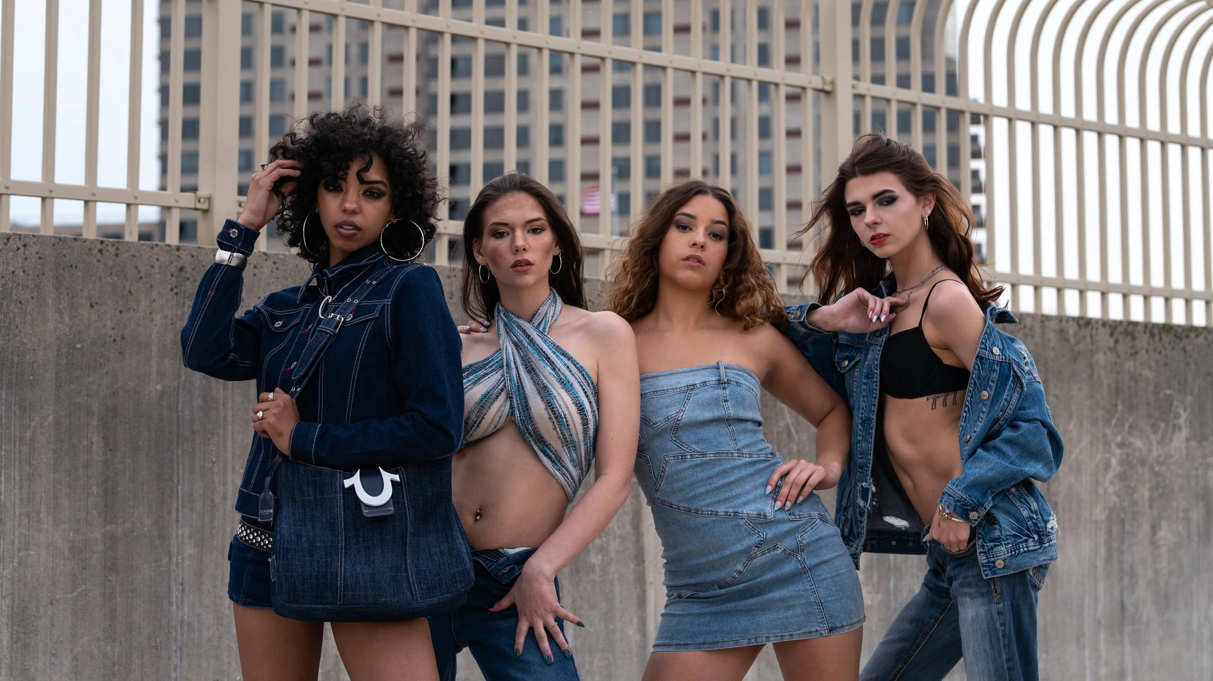 Four women posing in front of a concrete wall with a cityscape in the background, dressed in street fashion denim outfits and accessories.